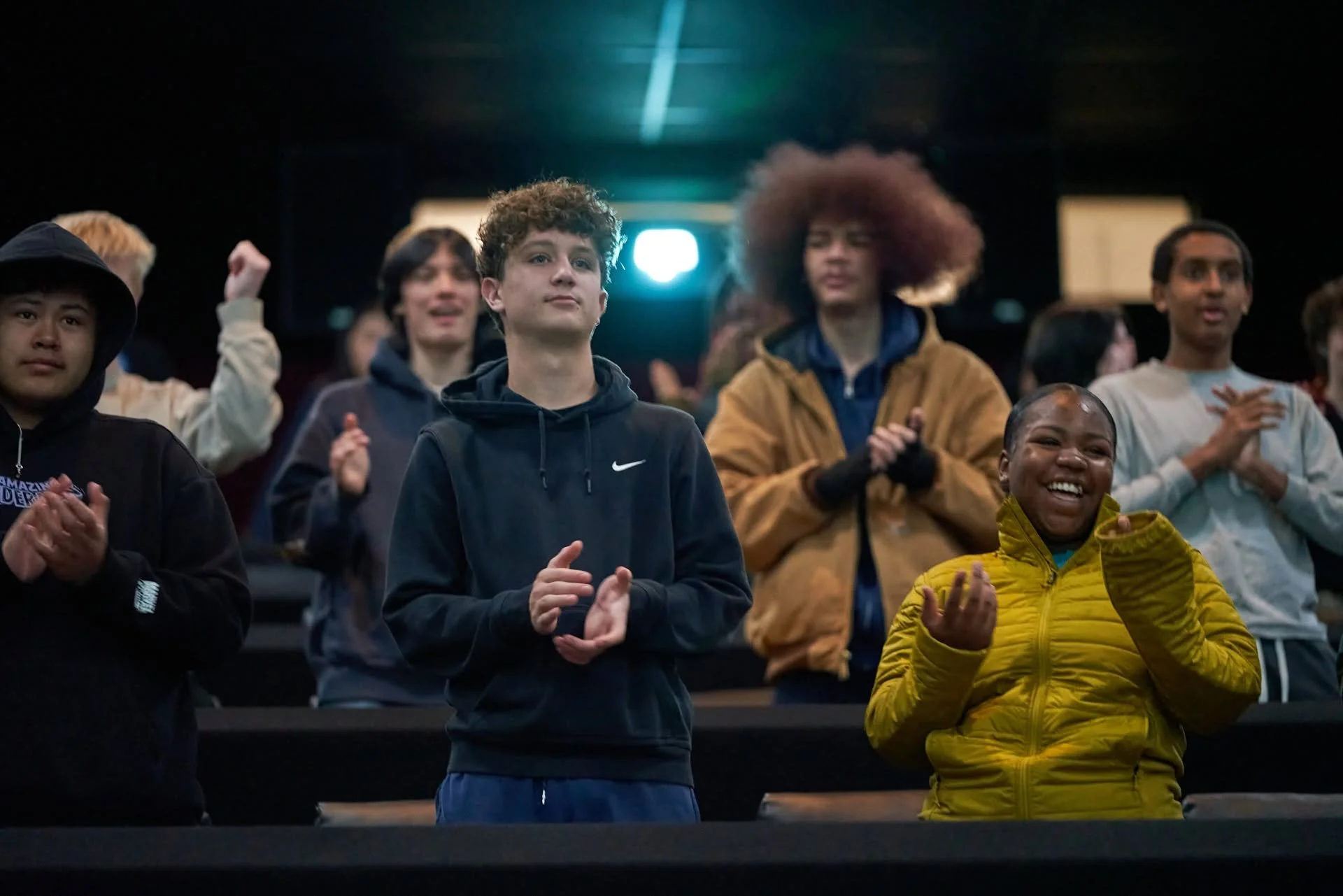 A group of young people standing in a dark auditorium, clapping and smiling, with some wearing jackets and hoodies.