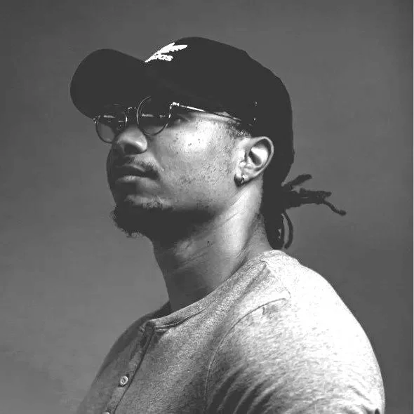A black and white photo of a man wearing glasses, a baseball cap, and a casual shirt, looking to the side.