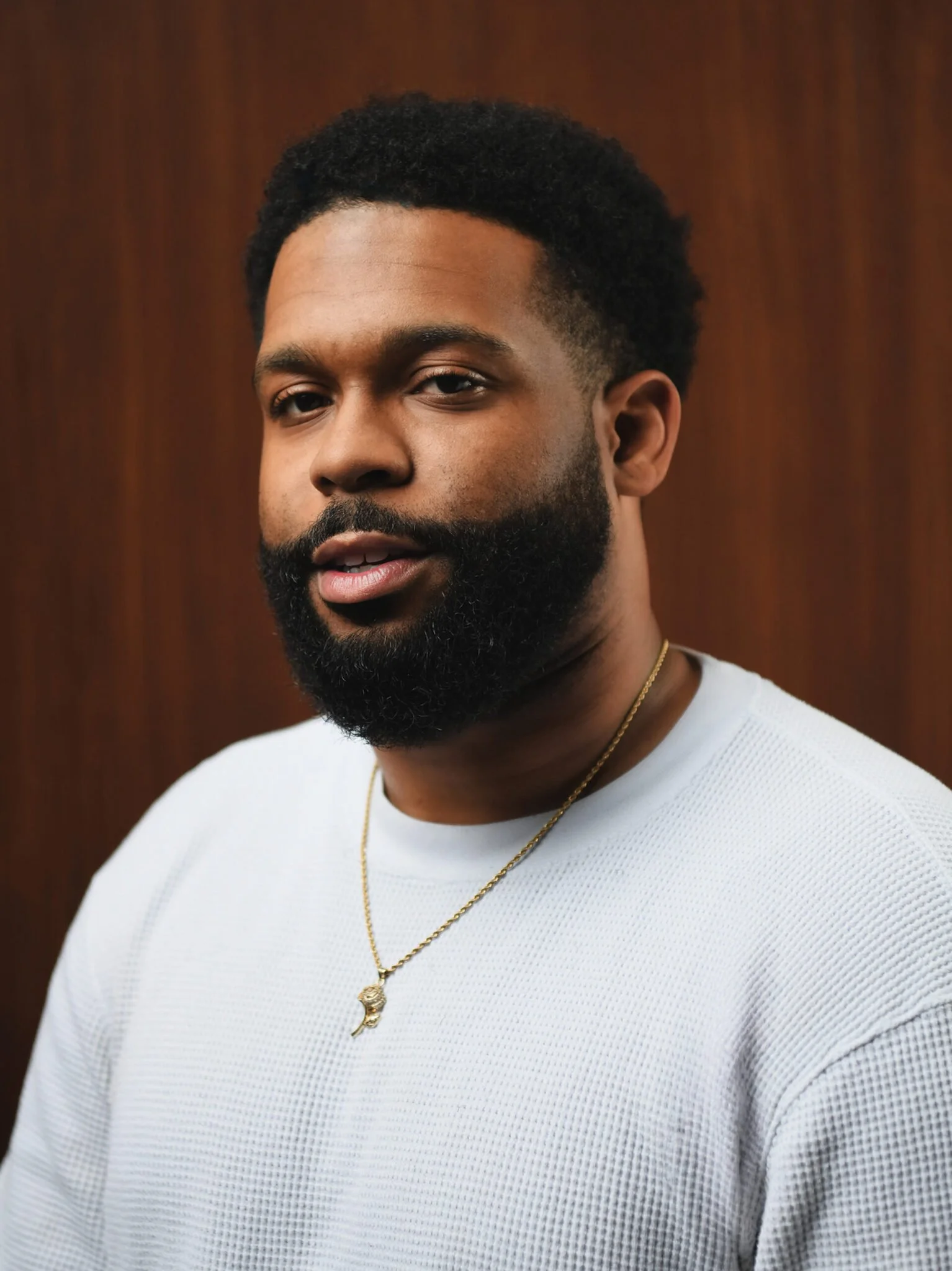 Close-up of a man with dark, curly hair, a full beard, wearing a white textured shirt, and gold chain necklace seen against a brown background.