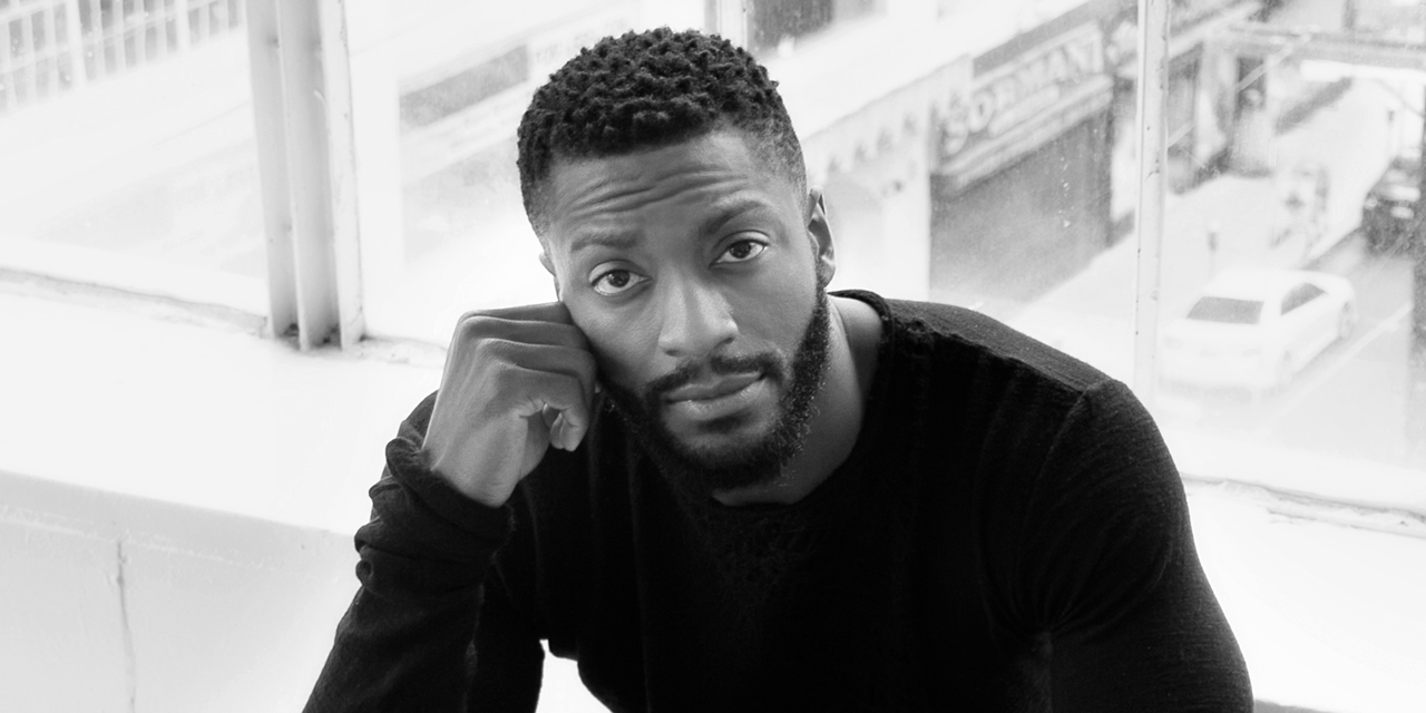 Black man with a beard, sitting by a window, resting his head on his hand, looking at the camera