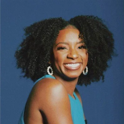 A woman smiling with curly black hair, wearing hoop earrings and a sleeveless blue top, against a blue background.