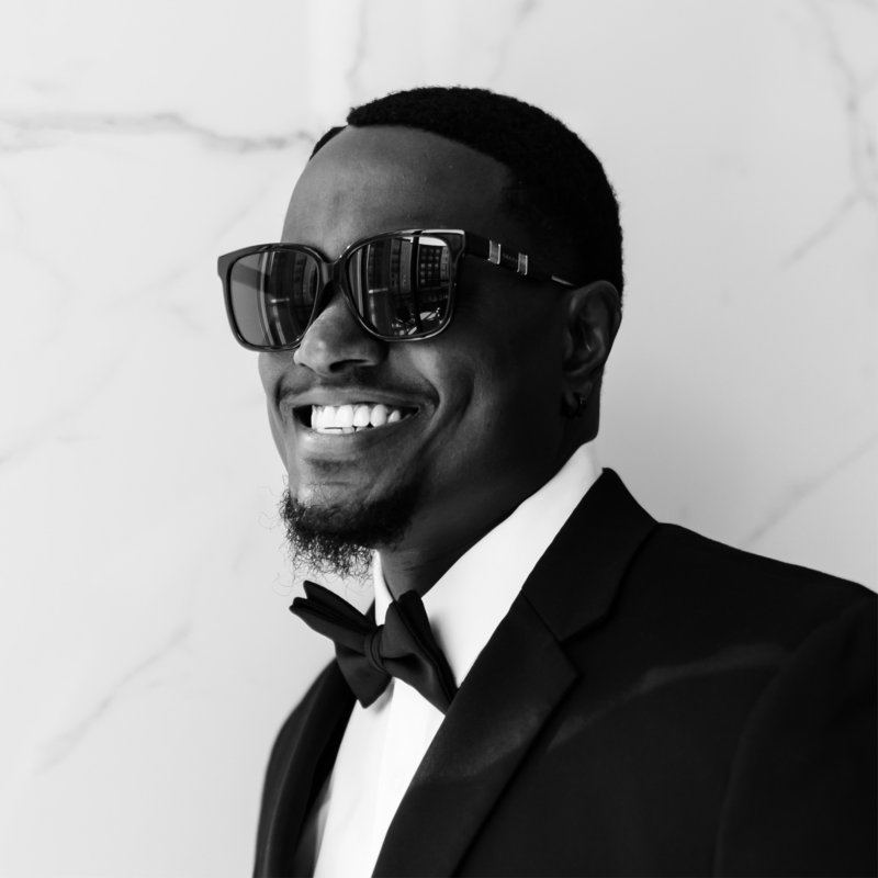 A man smiling with sunglasses, wearing a tuxedo and bow tie, in front of a light-colored marble wall.