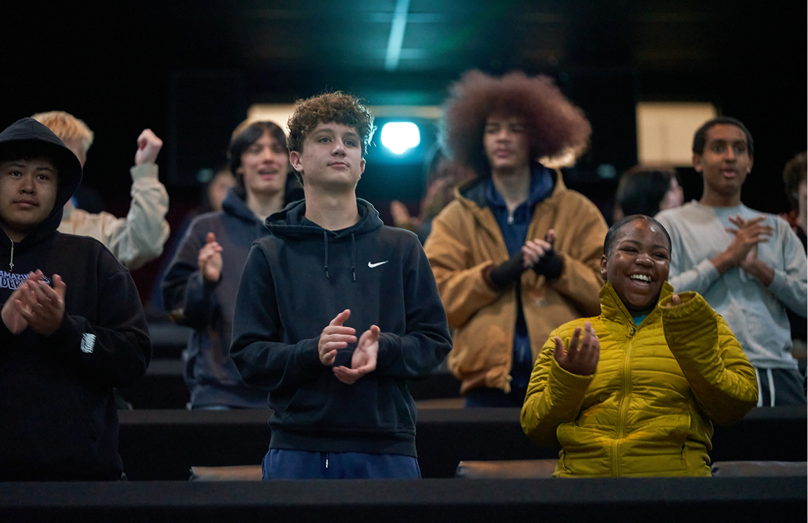 Group of people standing and clapping in a dark indoor setting, some smiling and others with neutral expressions.