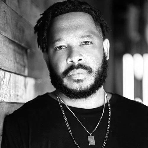 Black and white portrait of a man with a beard and dreadlocks, wearing a black shirt and layered necklaces, standing beside a wooden wall.