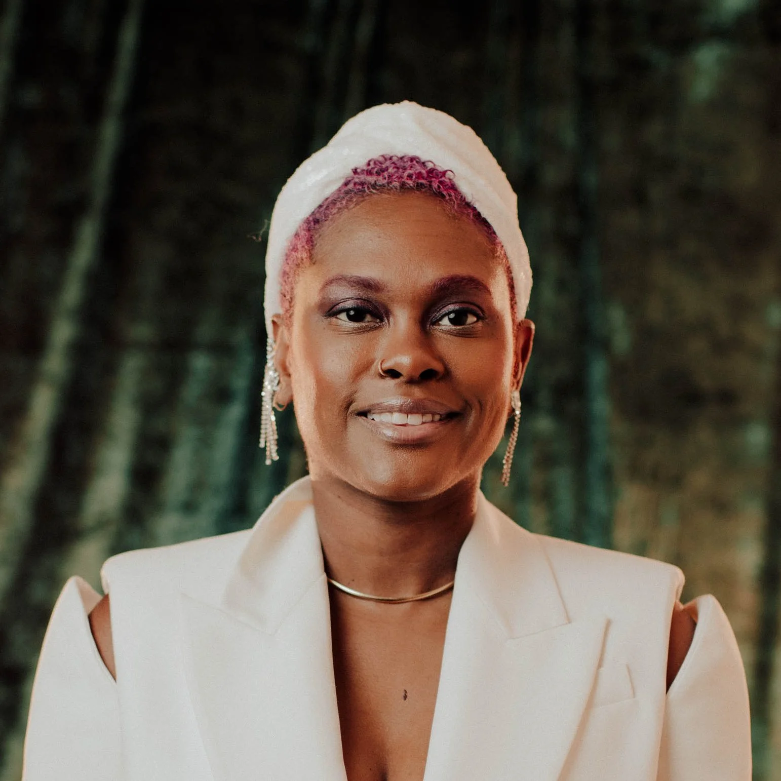 A woman with short pink curly hair, wearing a white headwrap, earrings, a necklace, and a cream-colored blazer, standing outdoors in a lush green forest.