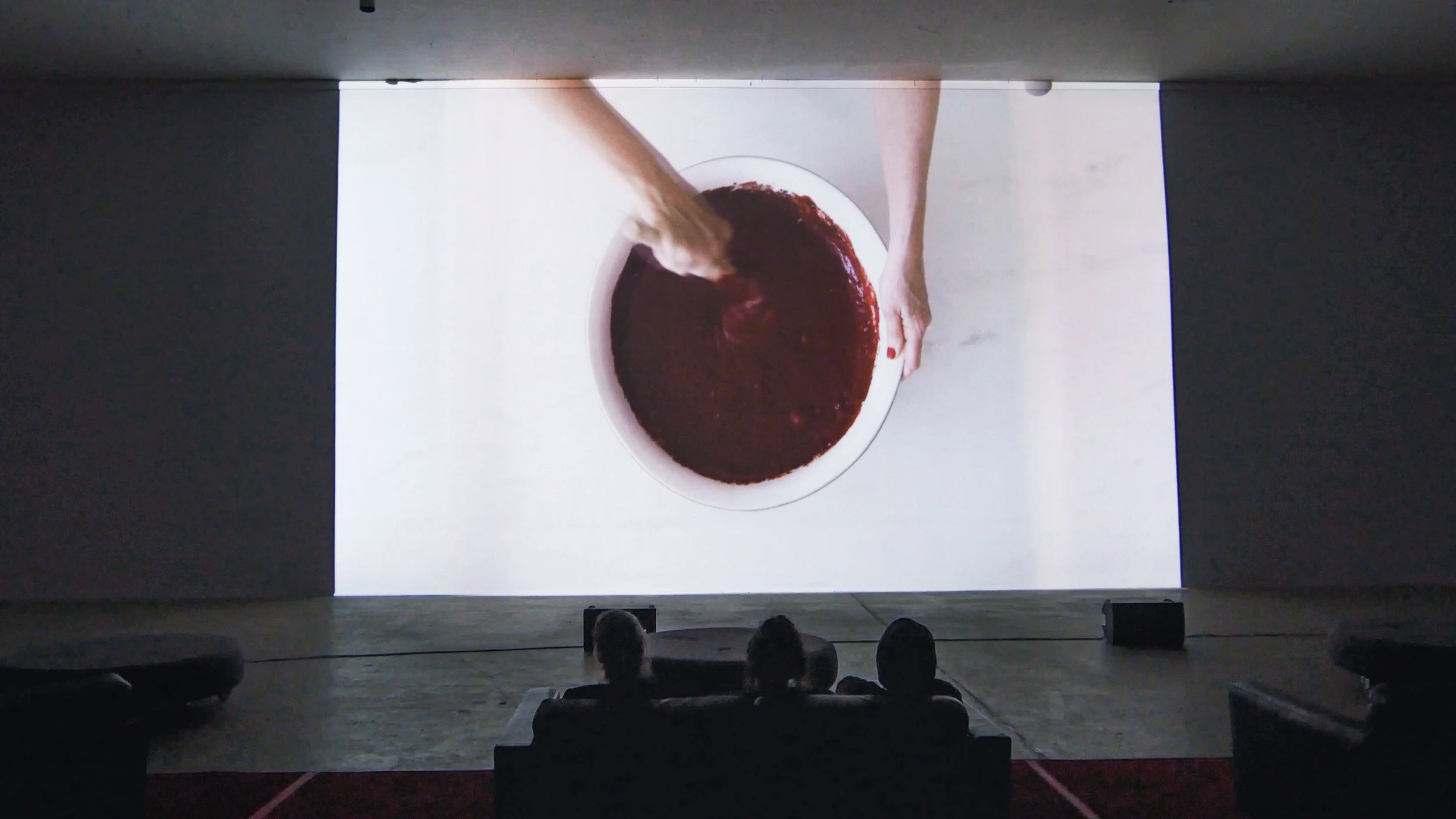 People seated in a dark theater watching a presentation that displays an overhead view of hands stirring a bowl of red liquid on a white surface.