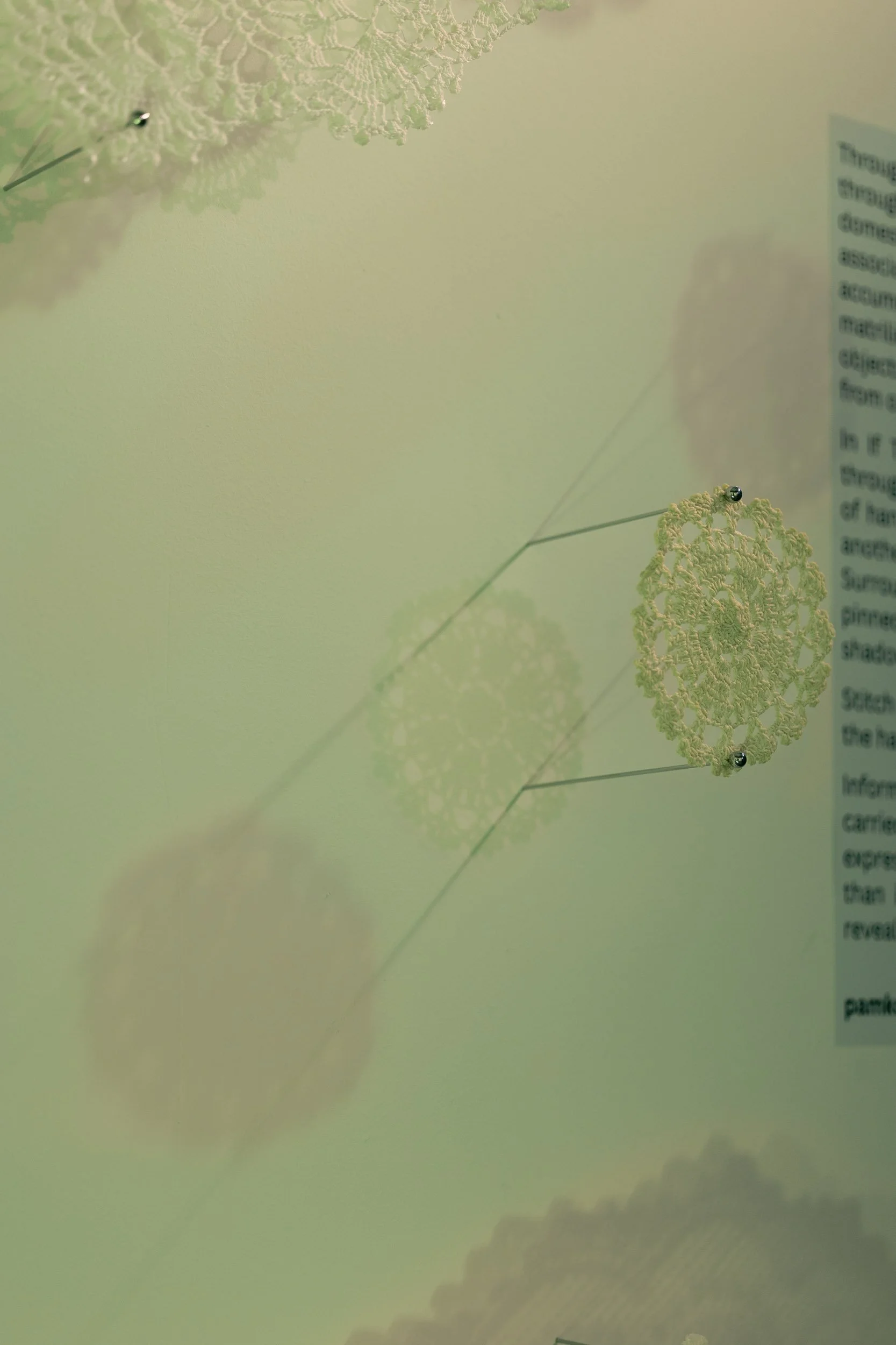 A single hand-crocheted doily on long pins hovering off the wall like a specimen under green stage lights that cast multiple echoes of magenta shadows.