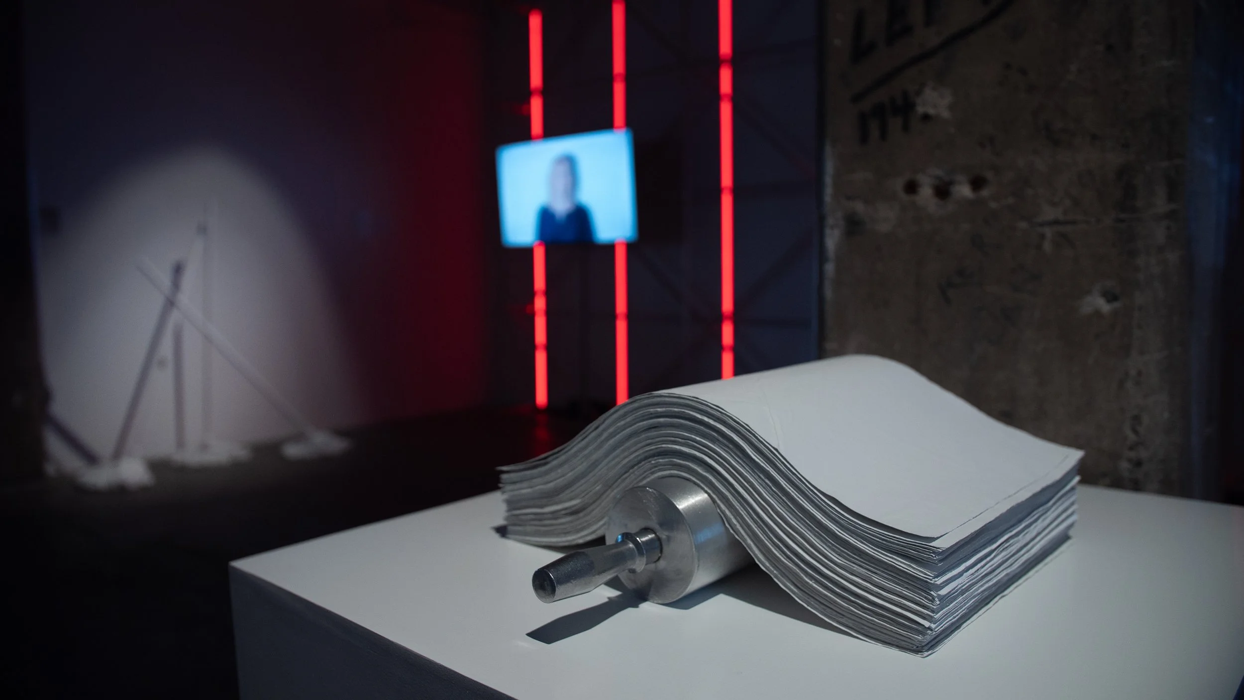 A paper-like sculptural material layered on aluminum rolling pin sculpture, and a blurred television screen in the background showing experimental video Screen Test with grandmother and mop sculptures.