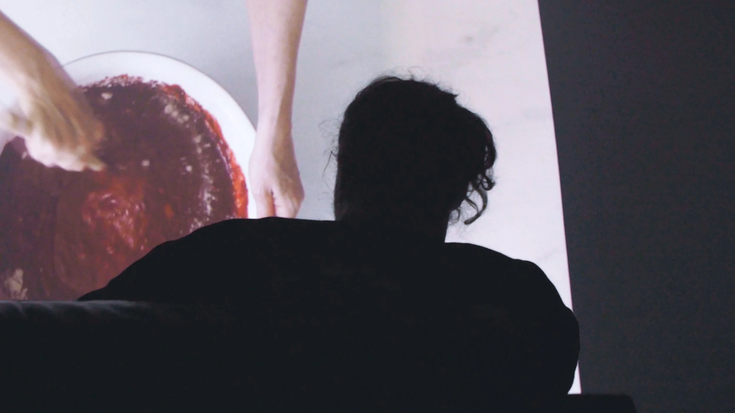 A person with curly hair, seen from behind, sitting and watching a large screen playing experimental video still 'SHE' at Icebox Project Space.