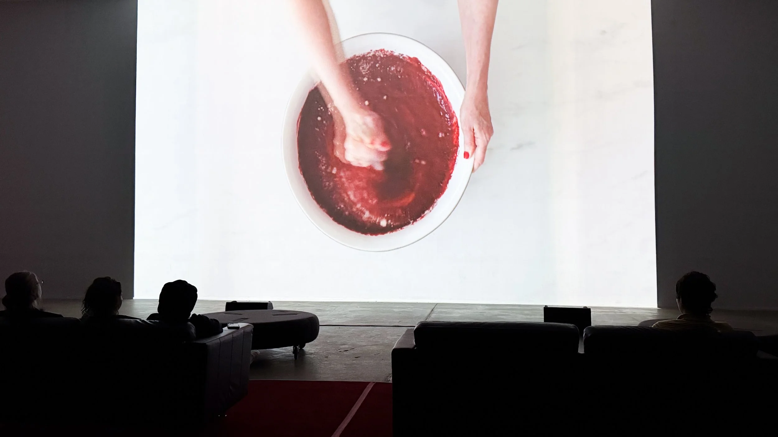 Experimental video still 'SHE' at Icebox Project Space, a woman repetitively mixing a blood red velvet cake mixture in a white bowl on a white marble surface. In front of the screen, people are seated and watching Pam Korman's video media.