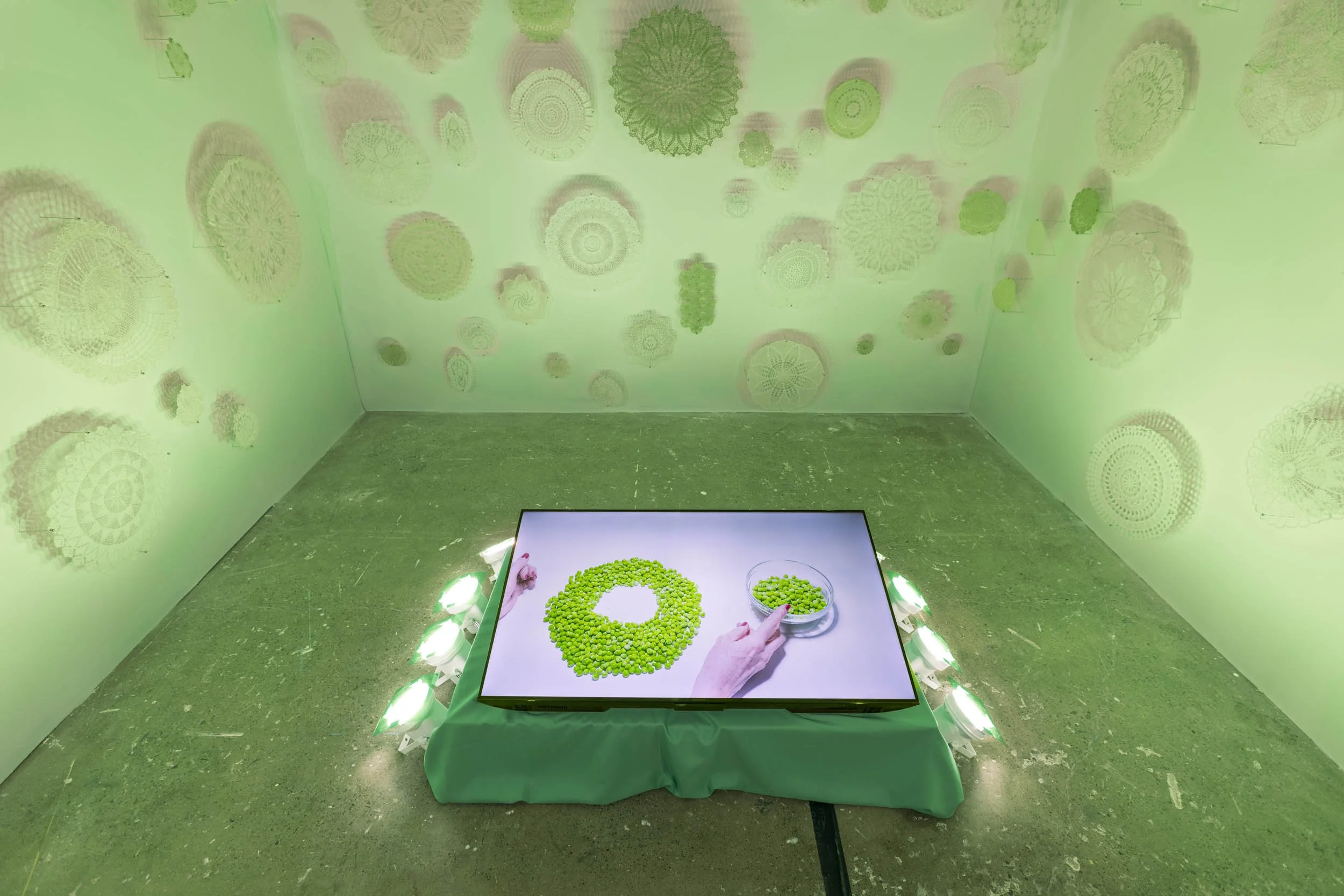 An art exhibition of hand-crocheted doilies under green stage lights with magenta shadows surrounding a green platform with a screen showing experimental video titled Peas.