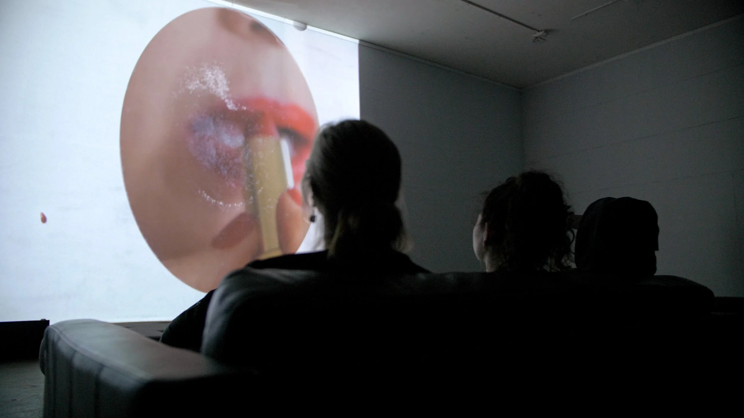 People seated in front of a large screen displaying an experimental video of a person's face with lips and nose close-up, at multimedia exhibit 20/92 Video Festival at Icebox Project Space.