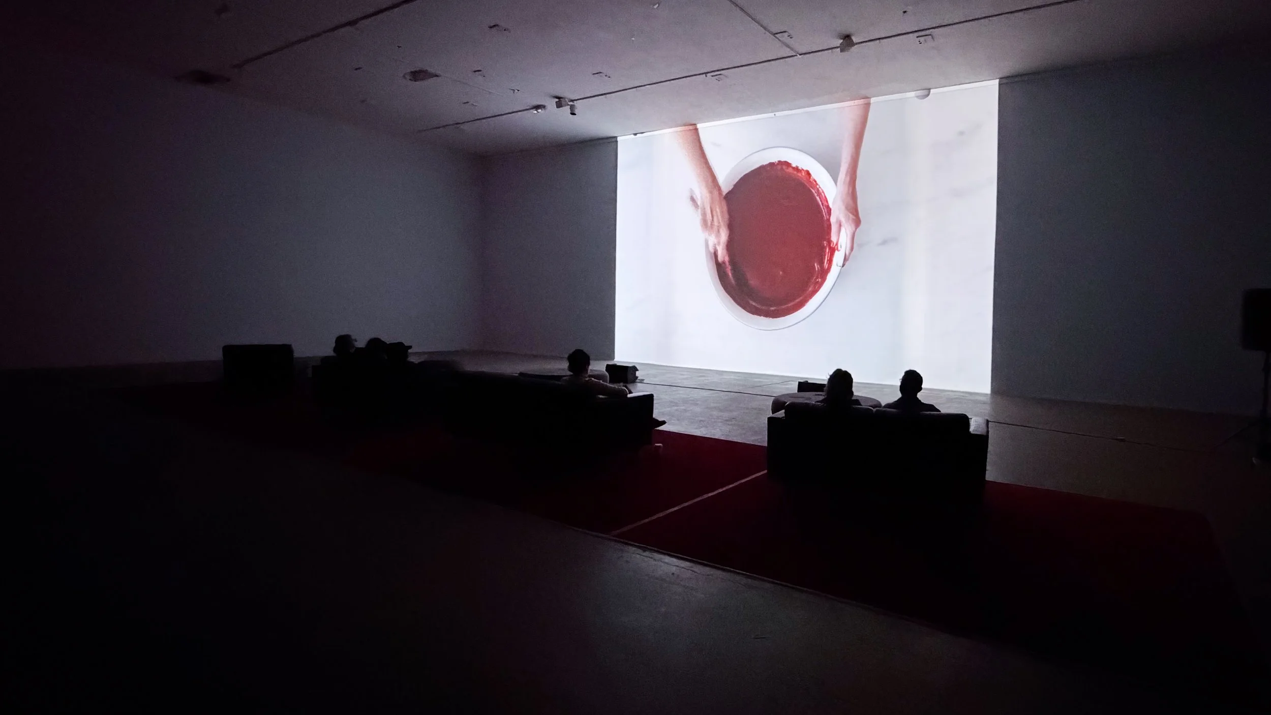 Dark room with a large screen displaying experimental video still 'SHE' at Icebox Project Space, a woman repetitively mixing a blood red velvet cake mixture in a white bowl on a white marble surface.