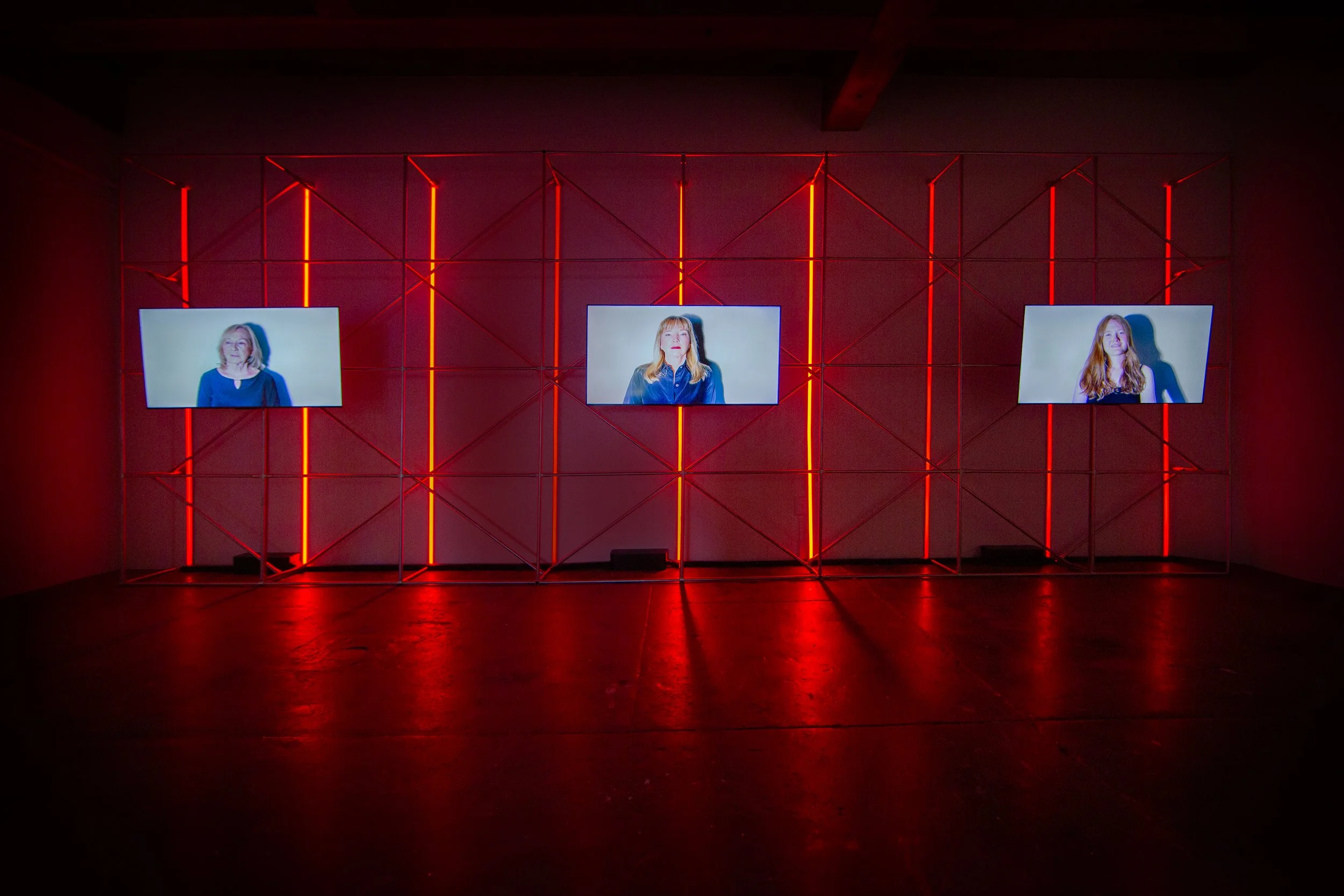 Three framed photographs of women are displayed on a metal grid structure against a dark background illuminated by red neon lights.