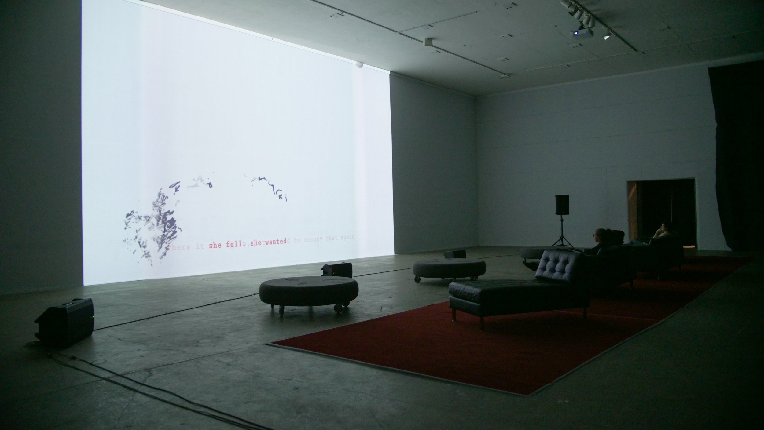 A large, dimly lit room with a high ceiling and a white projection screen displaying text from Korman's 'SHE'. Two people are seated, watching 20/92 Video Festival. 