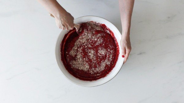 Two hands mixing red fruit filling with grated cheese in a white bowl on a light-colored surface.