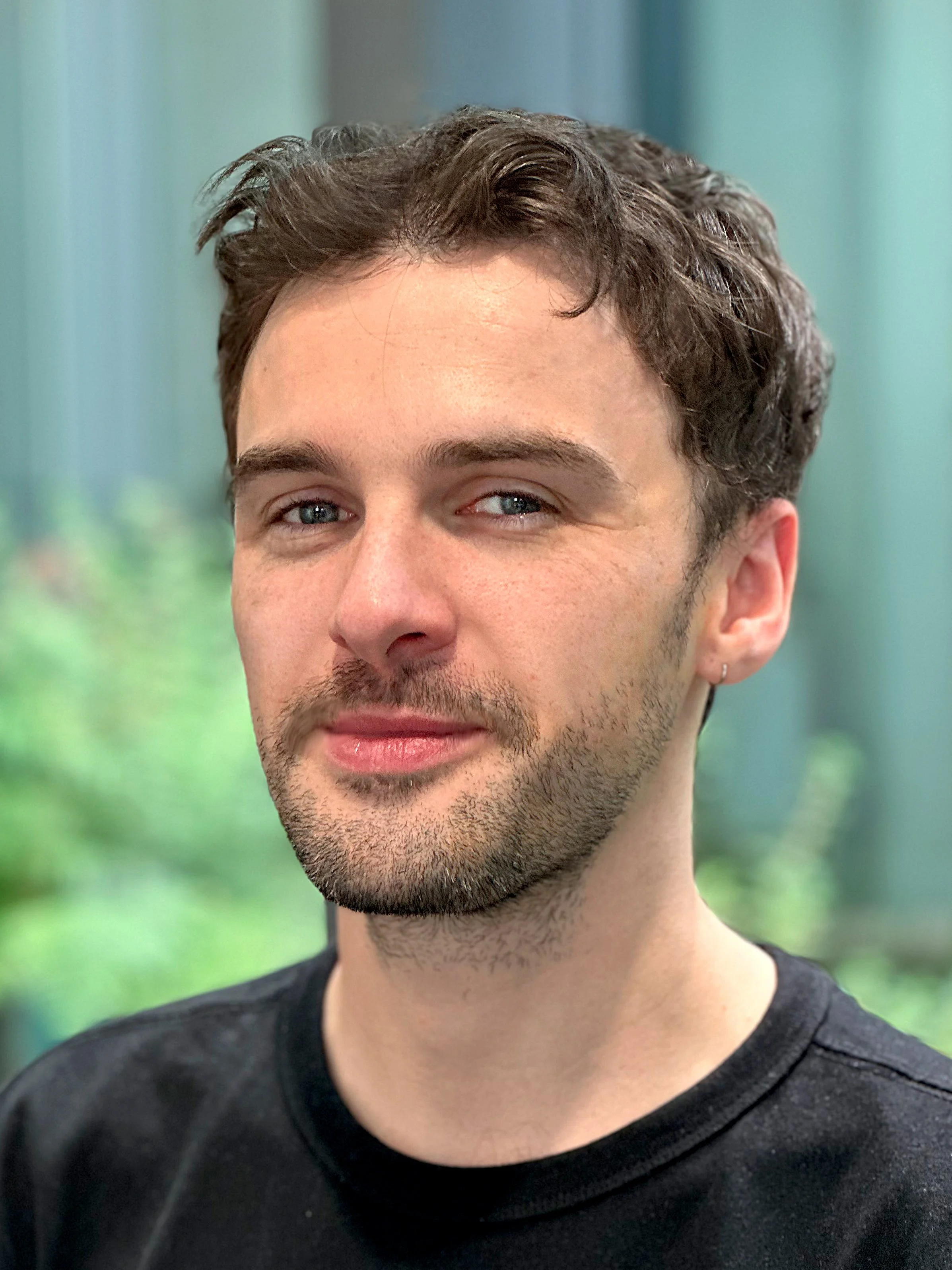 Close-up of a young man with brown wavy hair, light facial hair, and blue eyes, wearing a black shirt, with a blurred green and blue background.