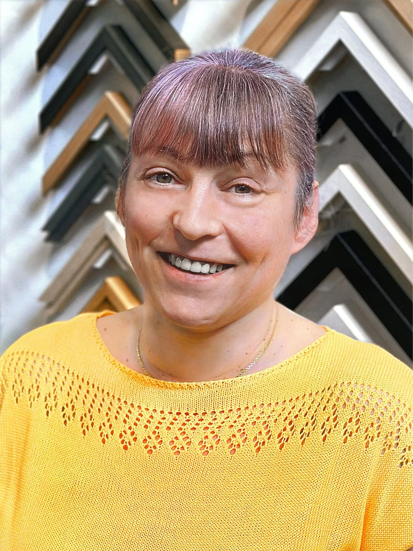A woman with red hair and tattoos on her left arm, wearing a blue sleeveless top, standing in front of a wall decorated with colorful picture frame corners arranged in a pattern.