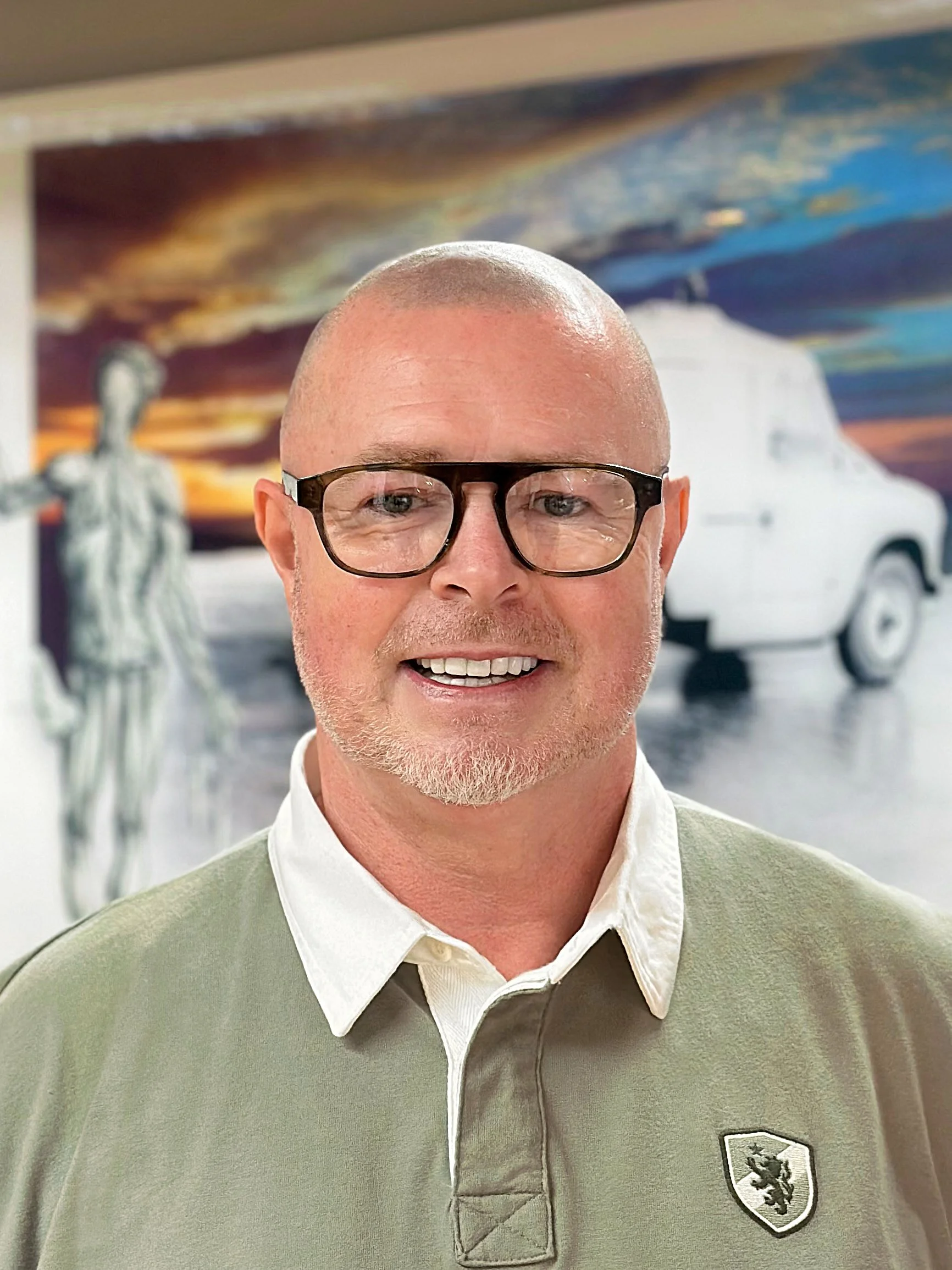 A smiling man with a bald head and a beard, wearing a plaid shirt, standing in front of a wall with framed artwork.