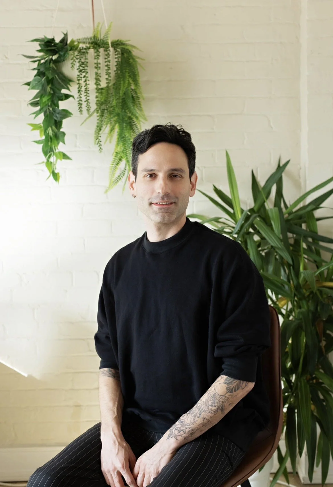 A man with dark hair, wearing a black shirt, sitting in front of a white brick wall decorated with green hanging plants and large leafy potted plants.