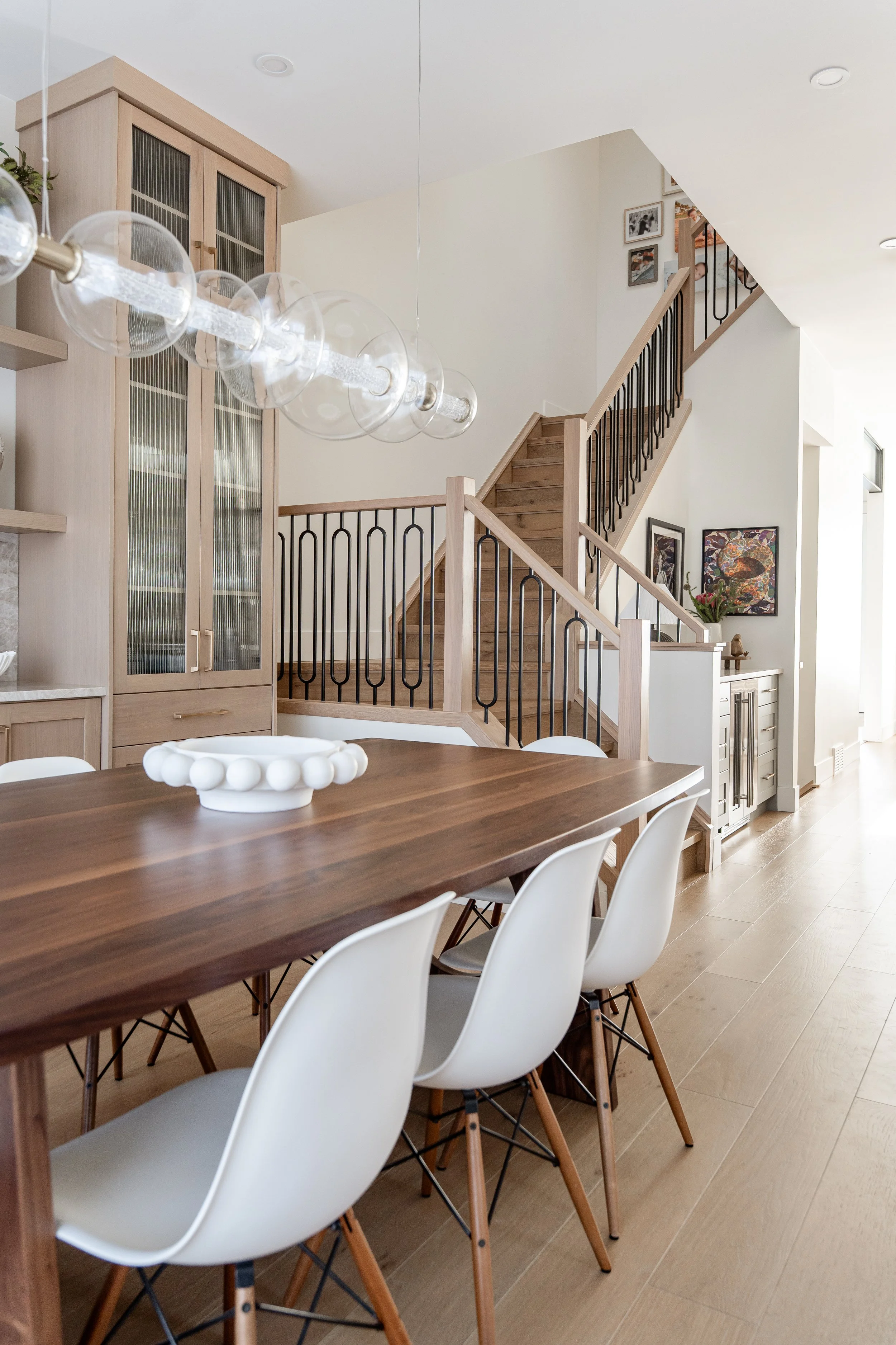 A dining table with white chairs, a white decorative bowl, a wooden staircase with a black railing, light wood cabinetry, framed artwork on the wall, and a modern hanging light fixture in a bright, minimalist home interior.