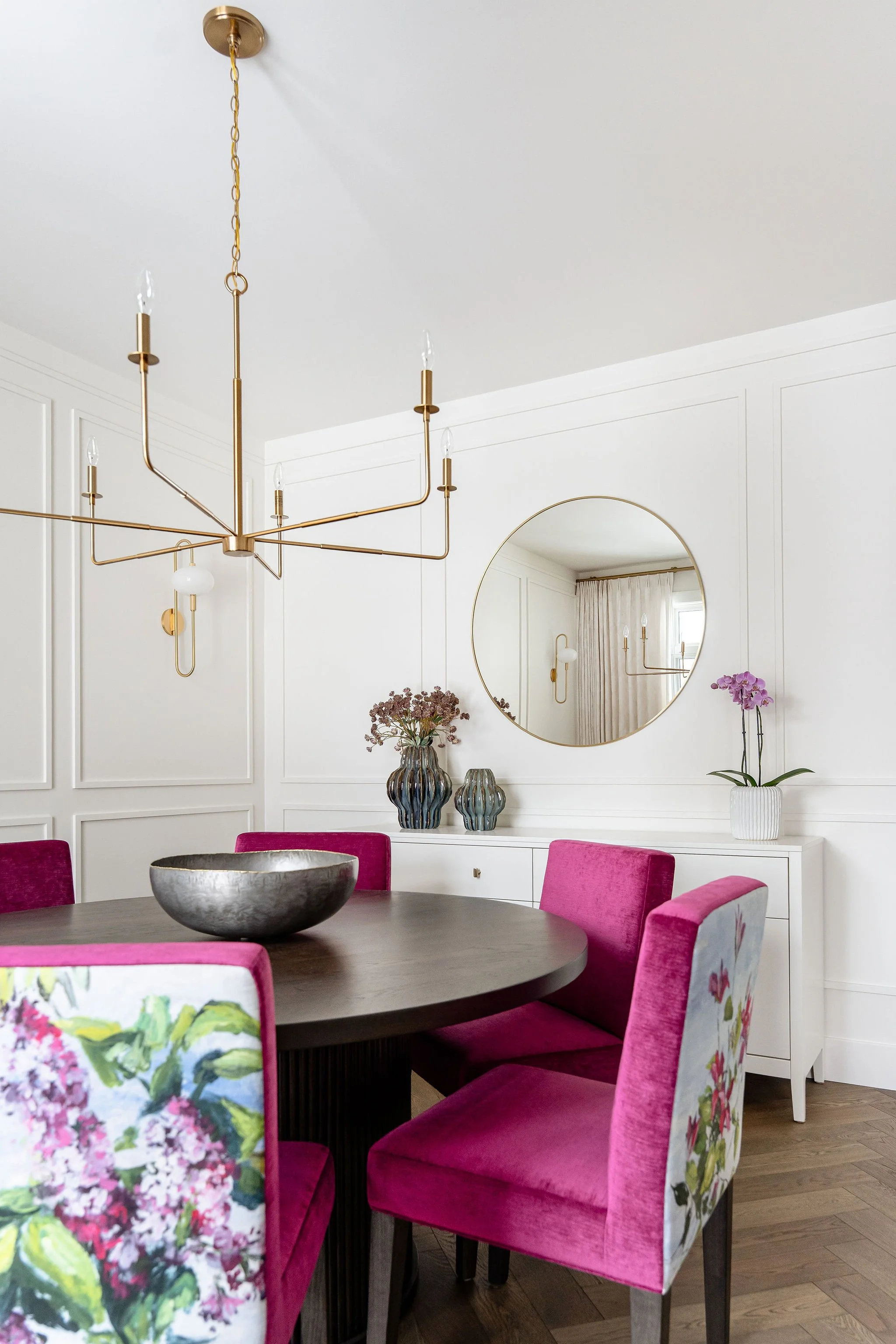A dining room with a round wooden table surrounded by six pink upholstered chairs, some with floral patterns on the back. On the table is a large metallic bowl. The background features white paneled walls, a large circular mirror, and decorative vase