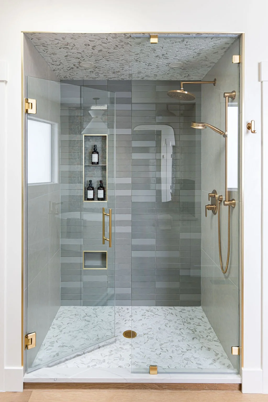 A modern glass-enclosed shower with a gold handle and fixtures, gray tiled walls, patterned ceiling, and built-in shower niche with bottles inside.