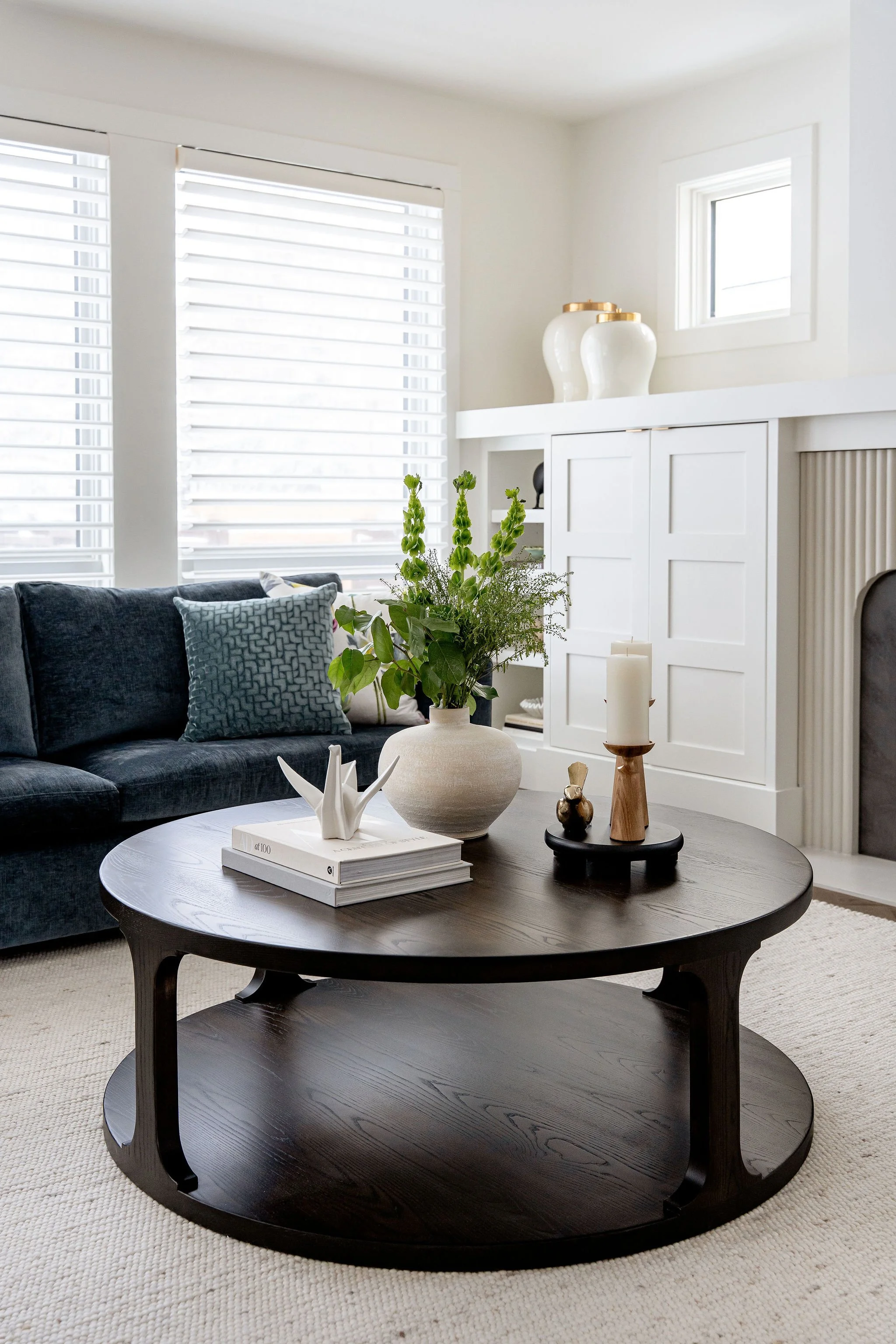 Living room with large windows, white walls, black sofa with patterned pillows, round dark wood coffee table with decorative items including a white sculpture, vases with green plants, candles, and a book, and a built-in white cabinet with white vase