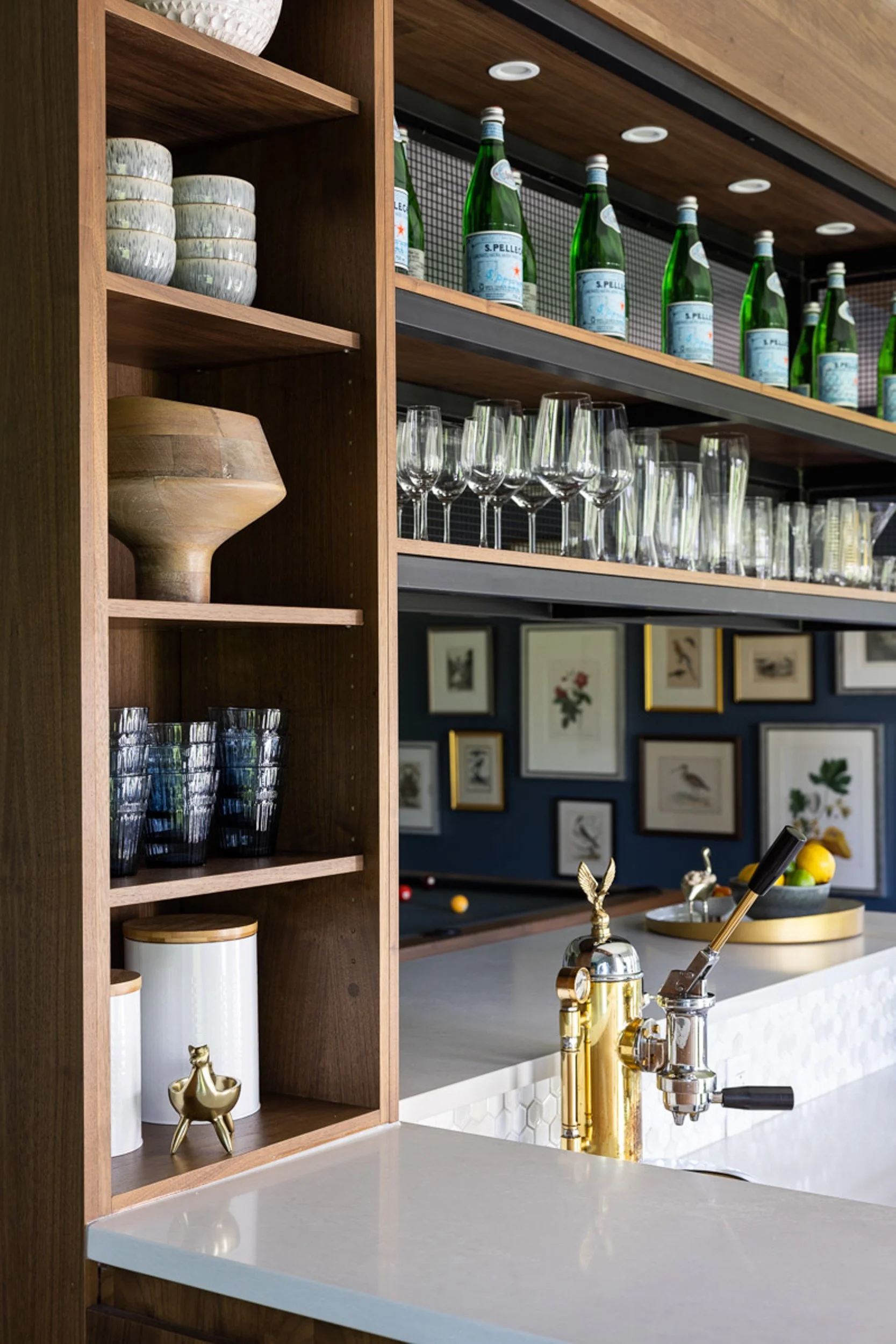 Modern kitchen with wooden shelves, glassware, bottled water, espresso machine, and framed artwork.