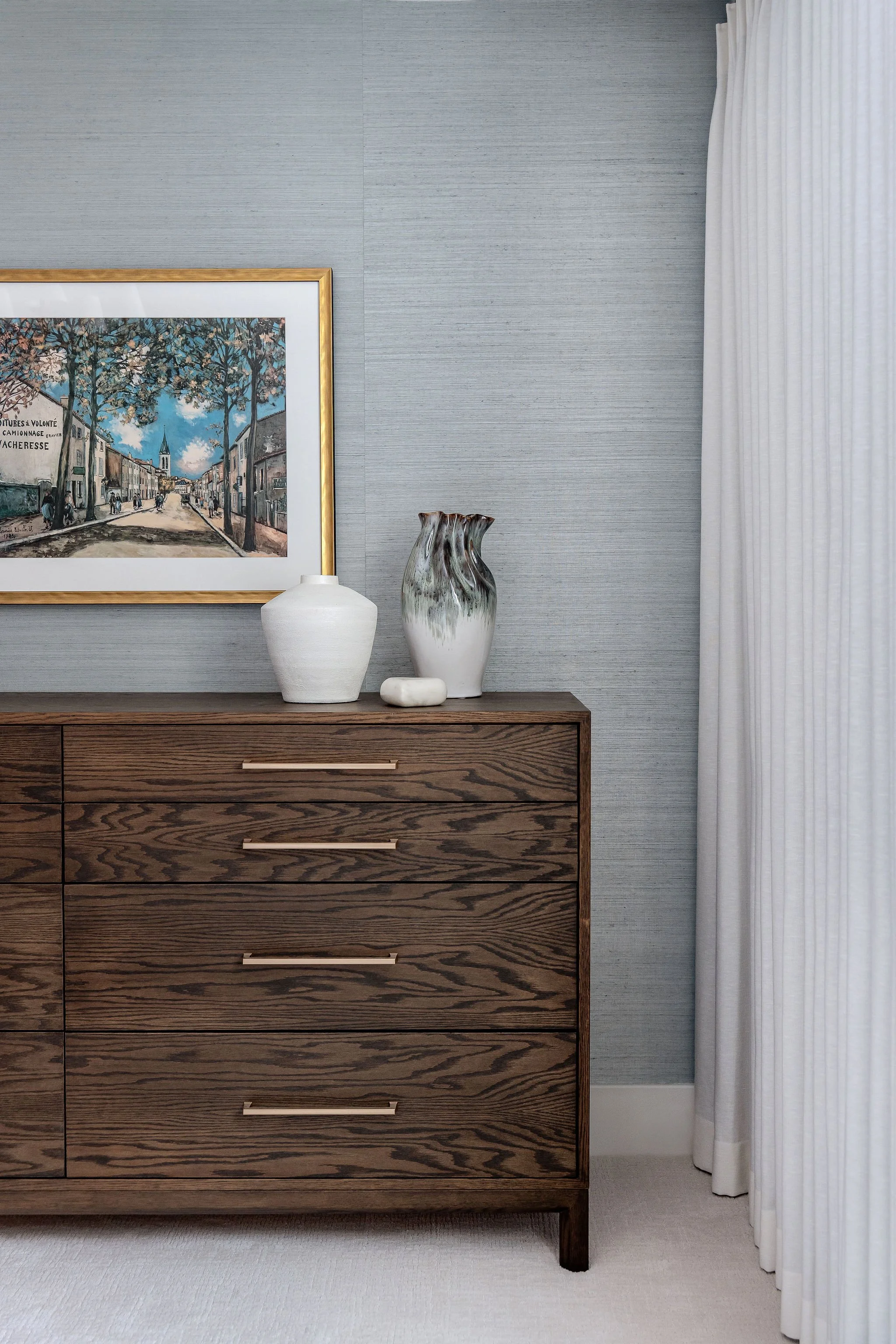 A wooden dresser with four drawers, decorated with three vases of varying sizes and a framed cityscape painting, is placed against a gray textured wall with white curtains on the right side.