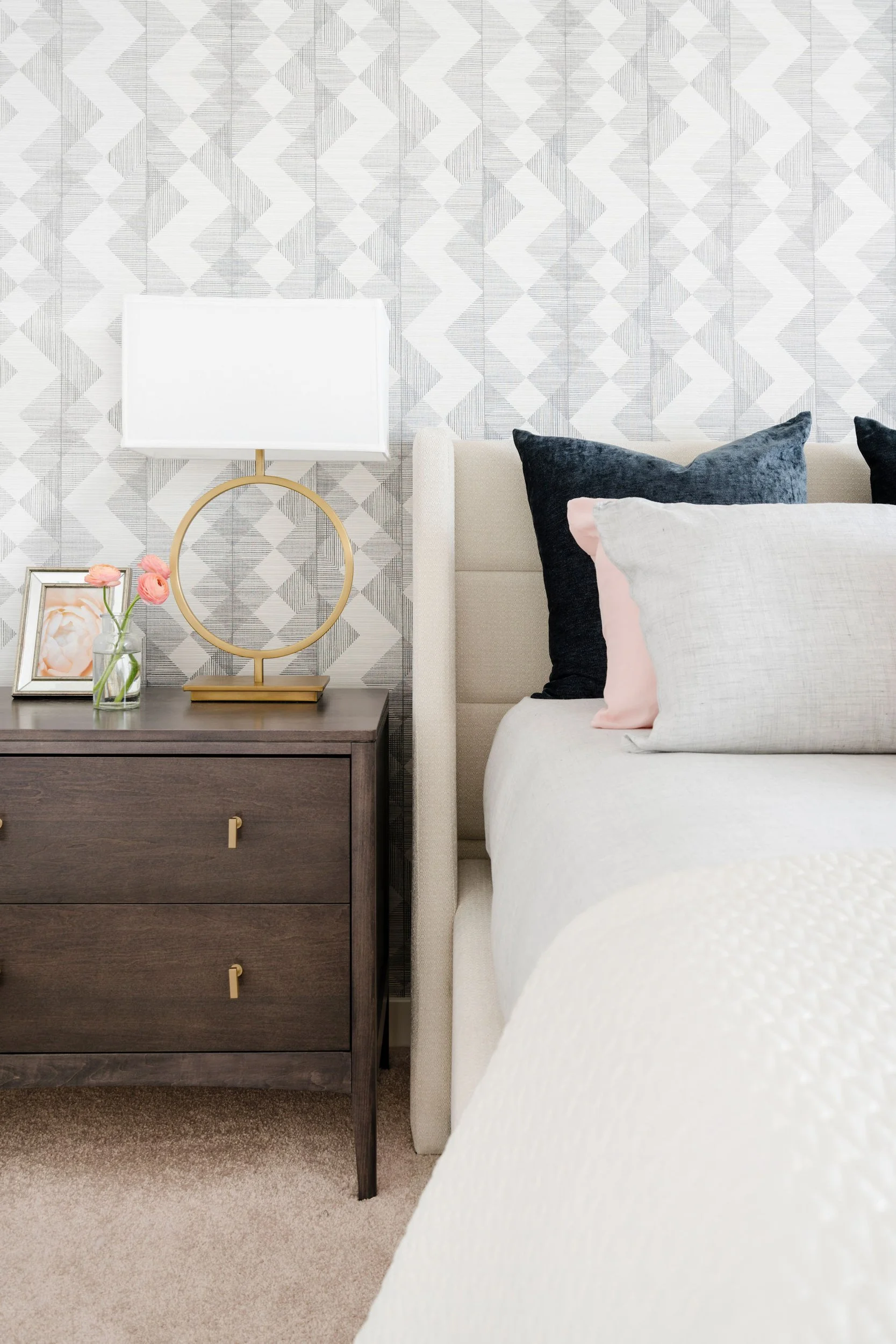 Modern bedroom with geometric wallpaper, a wooden nightstand, circular lamp, vase with pink flowers, framed photo, and a bed with dark and light pillows.