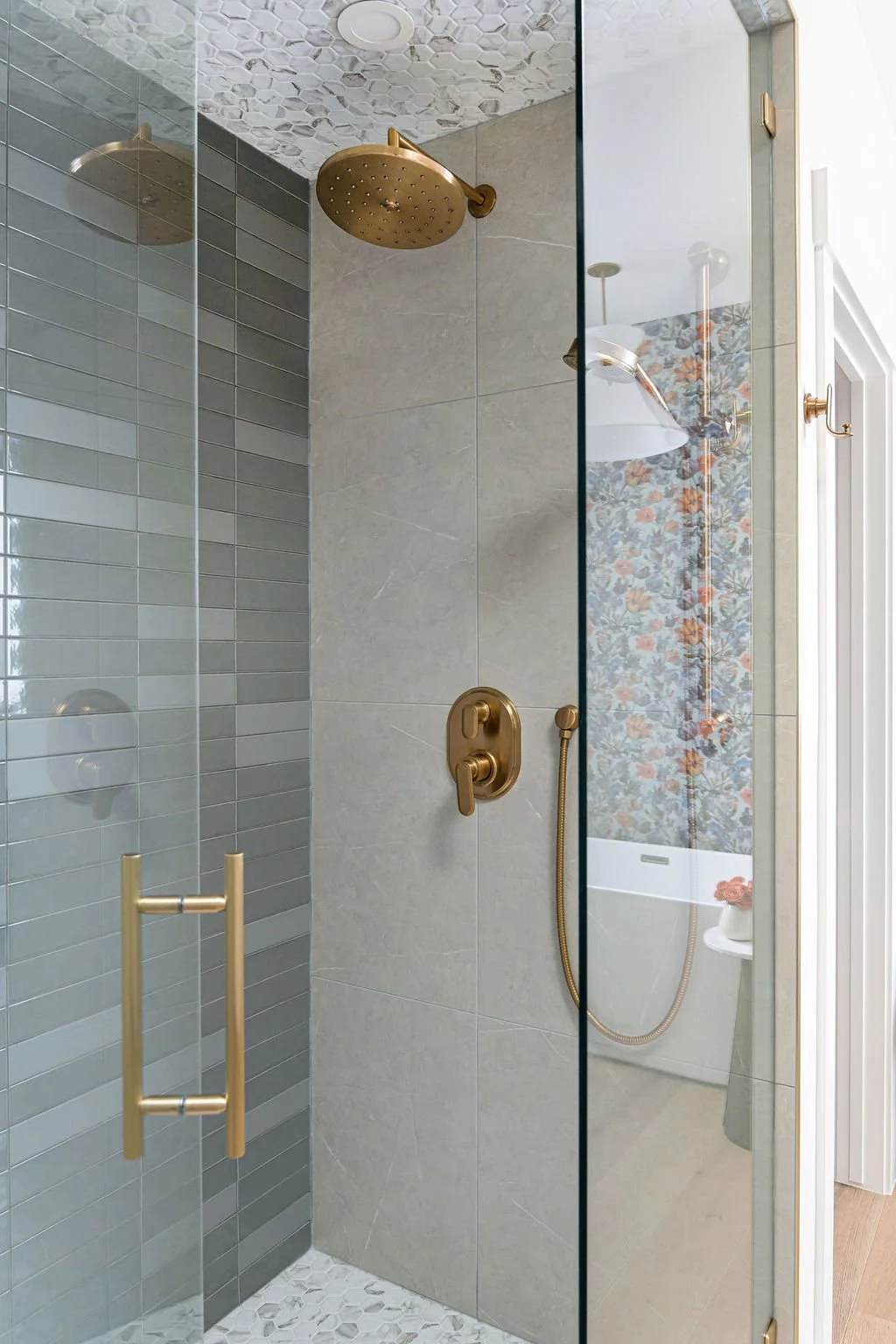 Modern walk-in shower with gold fixtures, including a rain showerhead and handheld spray, enclosed by clear glass doors, with textured and floral wallpaper reflection visible.