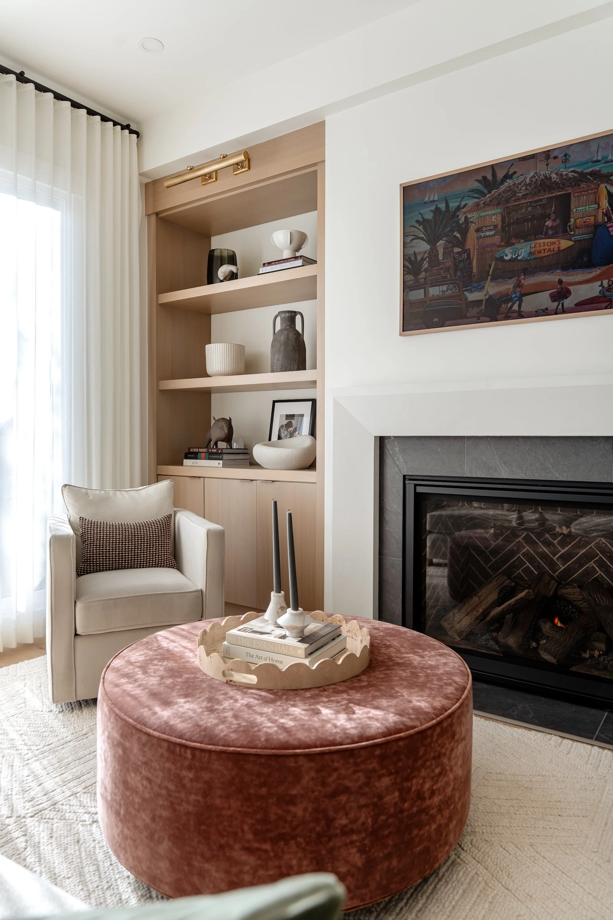 A cozy living room corner featuring a white armchair with a brown and white throw pillow, a round pink velvet ottoman with a tray of books and candles on top, a built-in wooden shelf with decorative objects, a fireplace, a framed artwork of a beach s