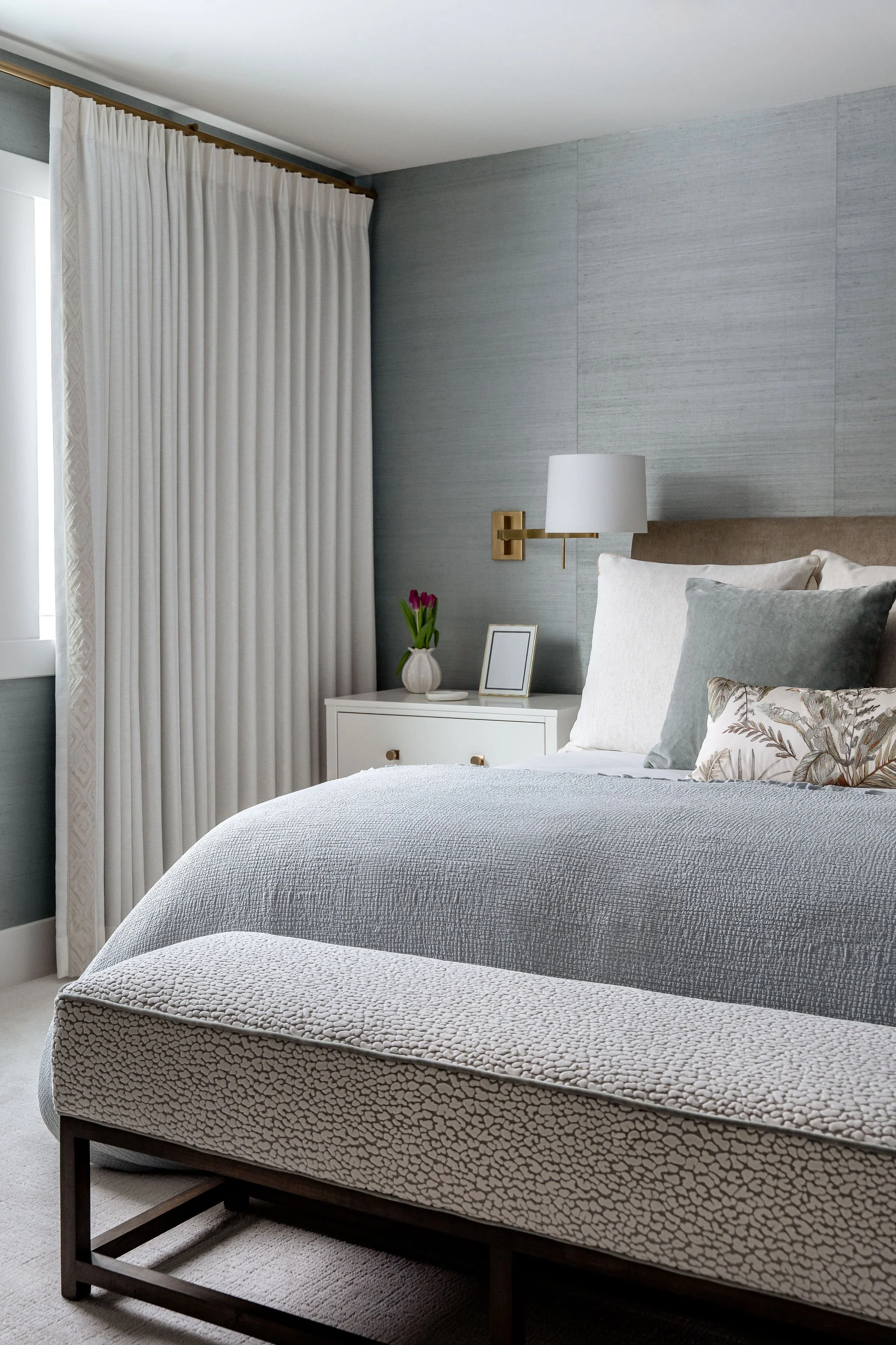 A bedroom featuring a neatly made bed with decorative pillows, a white bedside table holding a vase with pink tulips, a picture frame, gray textured wall, wall-mounted white lamp with brass accents, cream curtains, and a large window.