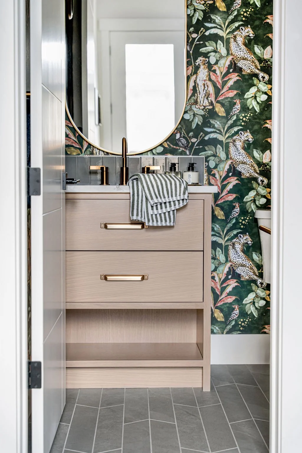 A bathroom vanity with a light wood finish, two drawers with gold handles, a striped towel hanging over the front, and a round mirror above. The wall behind features jungle-themed wallpaper with leopard, cheetah, tiger, and bird motifs. The vanity ha