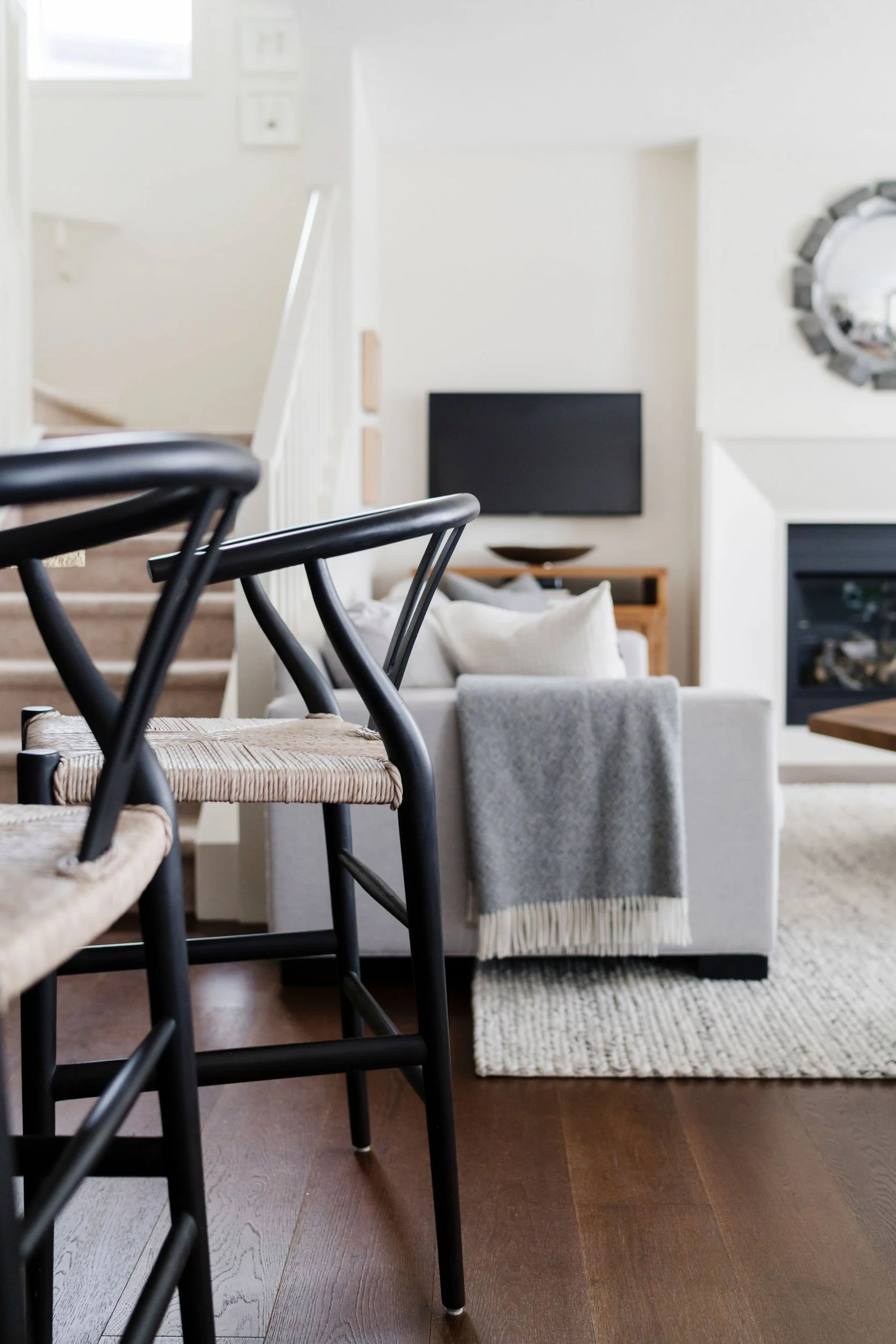 Modern living room with black bar stools, gray couch, and a wall-mounted TV. A throw blanket drapes over the couch, and a mirror hangs above the fireplace.