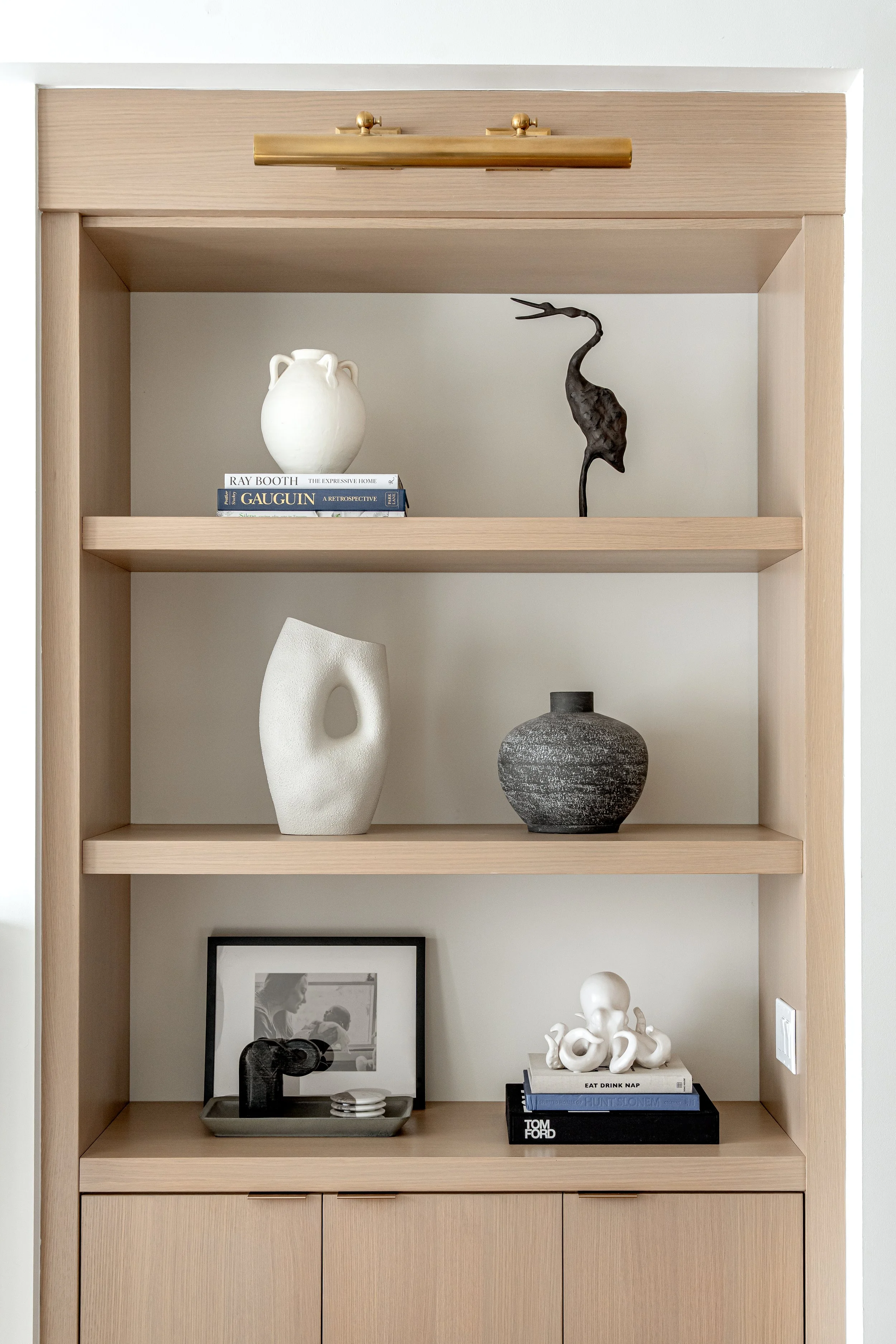 A wooden bookshelf with decorative items, books, and framed photos. The top shelf has a white ceramic vase and a black abstract sculpture of a heron. The middle shelf has a white oval-shaped vase, a black and white framed photo, and a tray with a bla