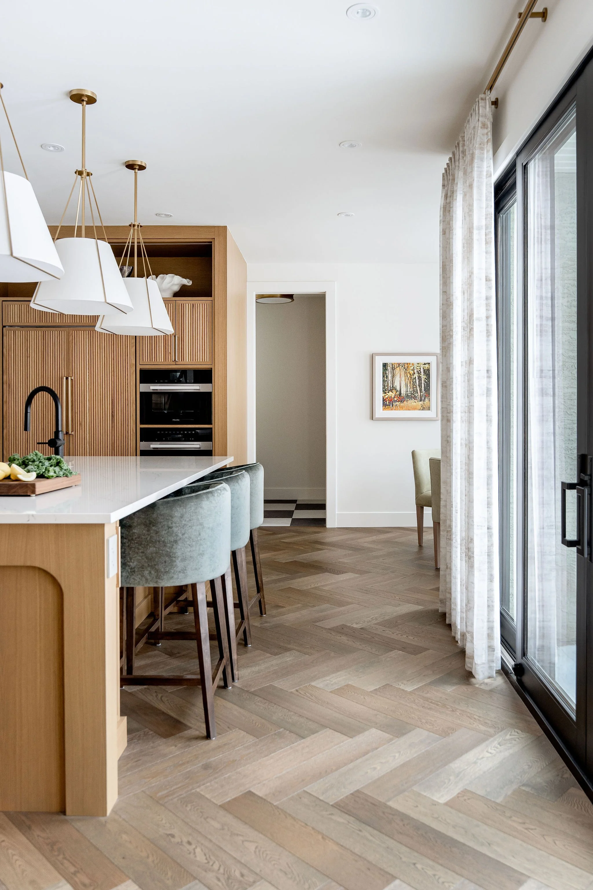 A modern kitchen with wooden cabinets, a white countertop, and a row of four light fixtures with white shades hanging from the ceiling. There is a black faucet at the sink and a cutting board with greens and lemons. A framed artwork hangs on the wall