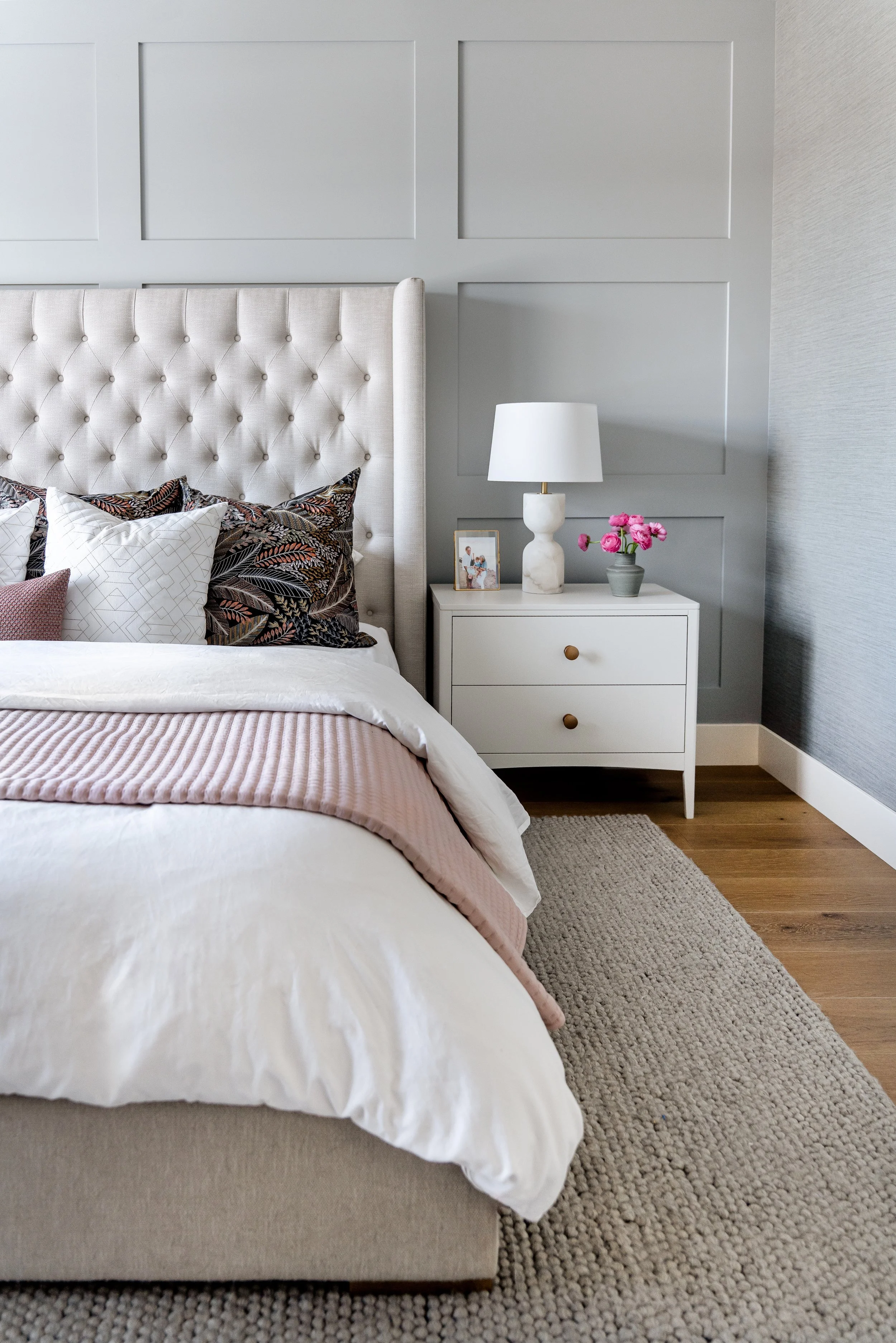 A bedroom with a beige upholstered bed with decorative pillows, a white nightstand with a lamp, framed photo, and pink flower arrangement, a textured beige rug, and paneled gray wall.