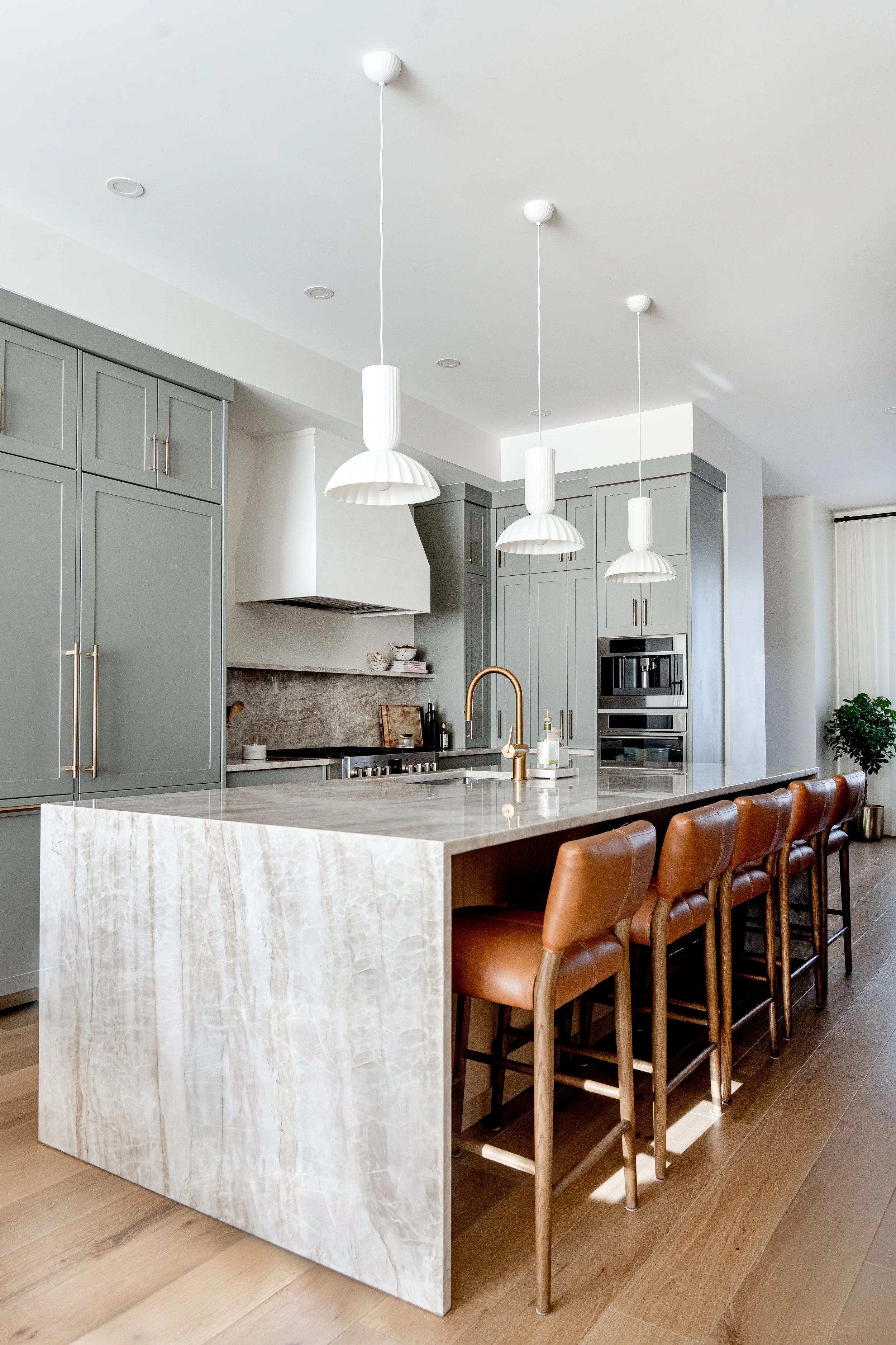 Modern kitchen with a large white marble island, gray cabinetry, three white pendant lights, and brown leather chairs at the island.