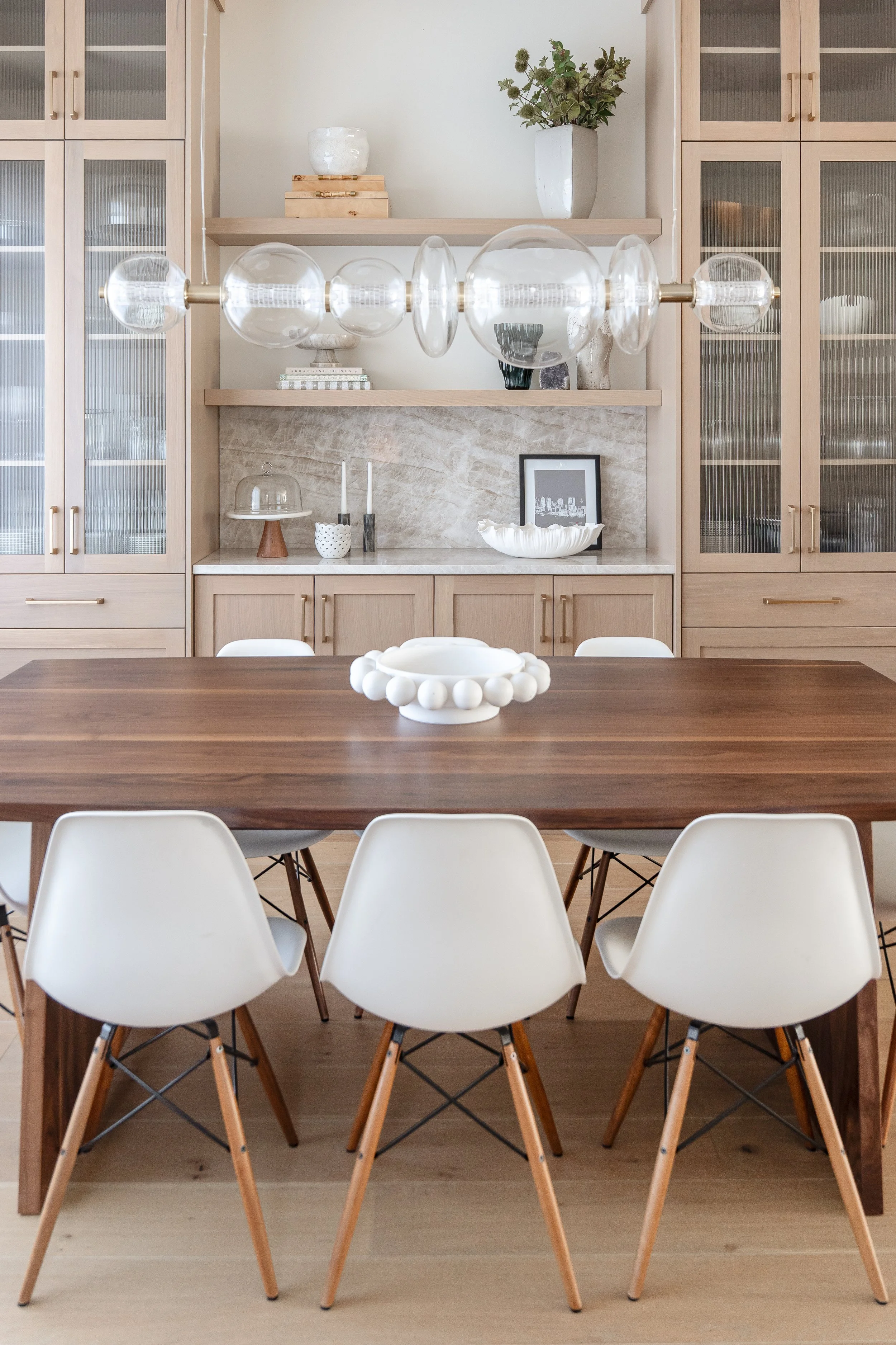 Modern dining room with a wooden table, white chairs with wooden legs, a built-in wooden cabinet with glass doors, and decorative items including a white bowl, candles, framed photo, and vases, with open shelves displaying a large glass sculpture and