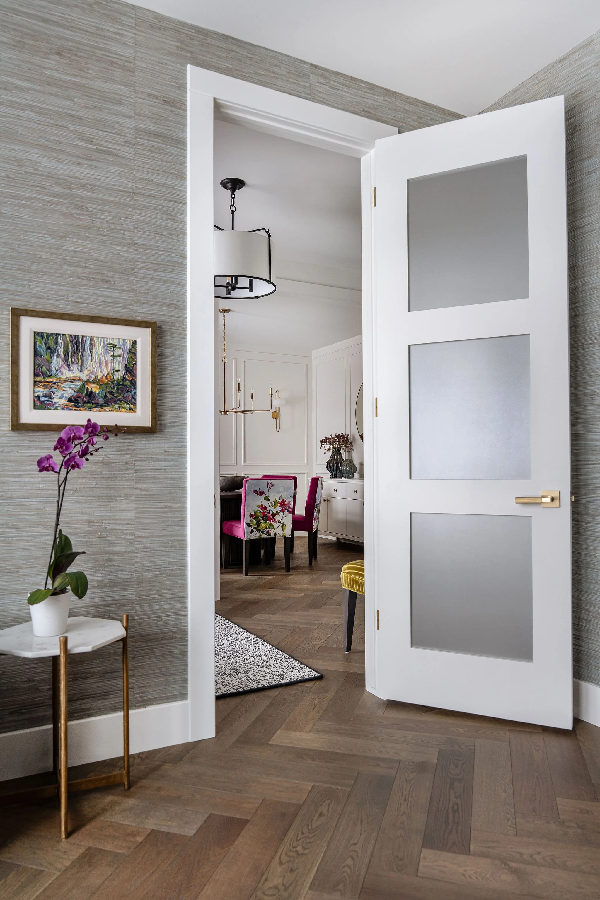 A home interior with frosted glass paneled white interior door open to a dining area with pink velvet chairs, white cabinetry, and a chandelier. A wall features a colorful landscape painting and a small side table with a white pot holding an orchid. 