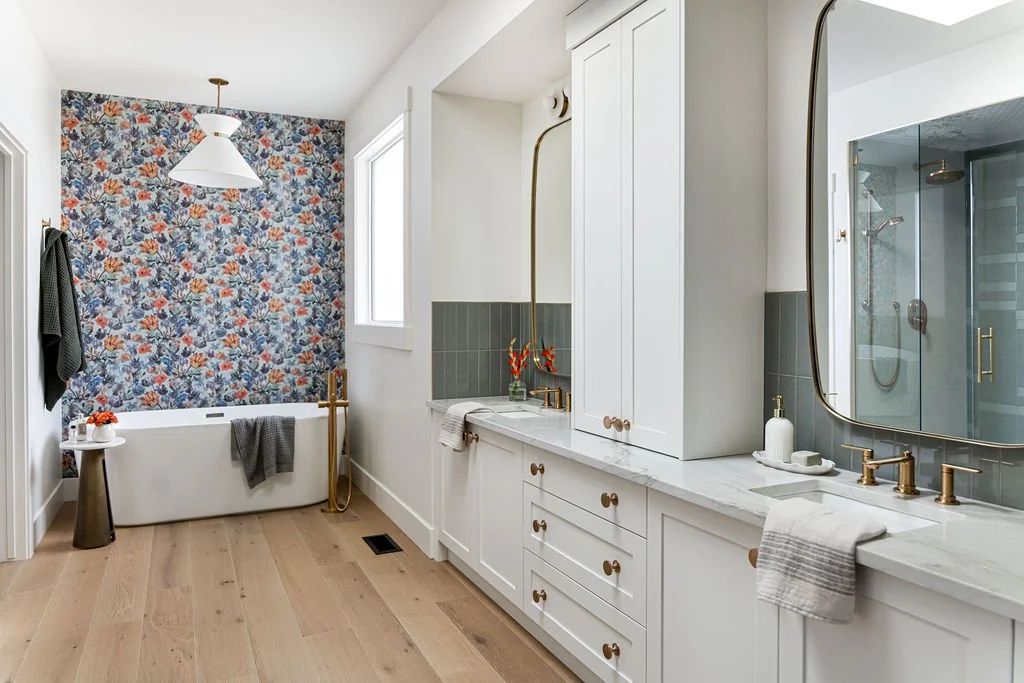 Modern bathroom with a floral accent wall, white vanity with gold fixtures, mirror, grey tiles, bathtub, and walk-in shower.