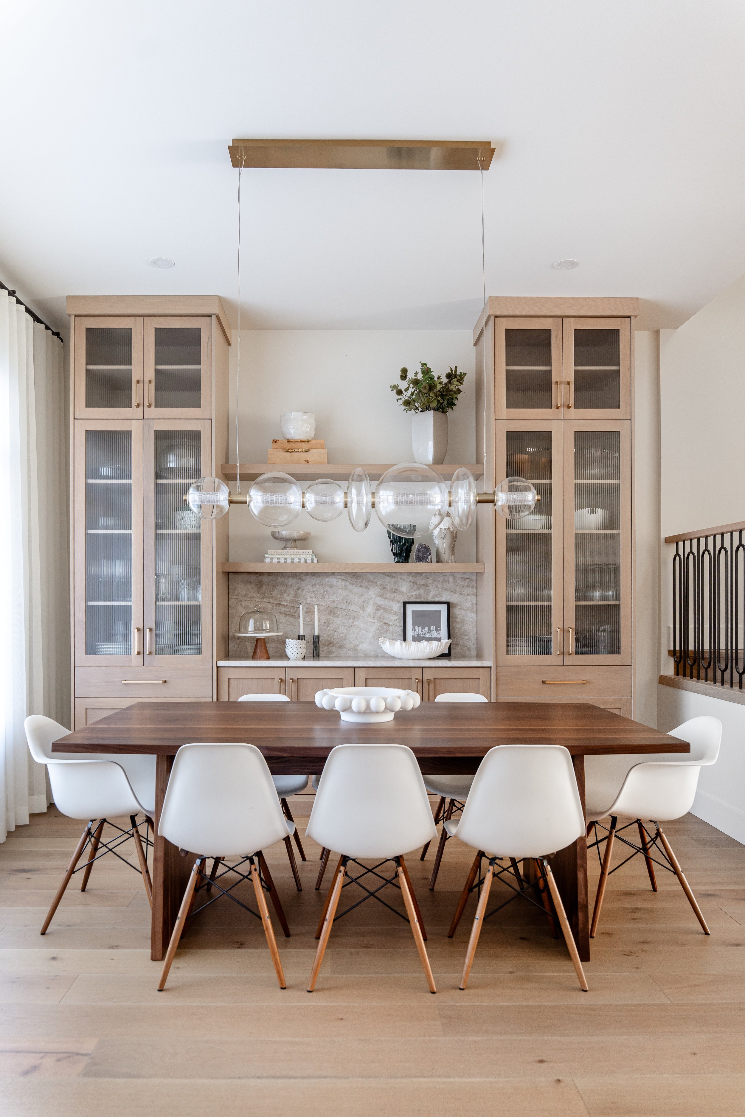 Modern dining room with a wooden table, white chairs, built-in cabinets with glass doors, a chandelier, and decorative items on the shelves including a vase and framed photo.