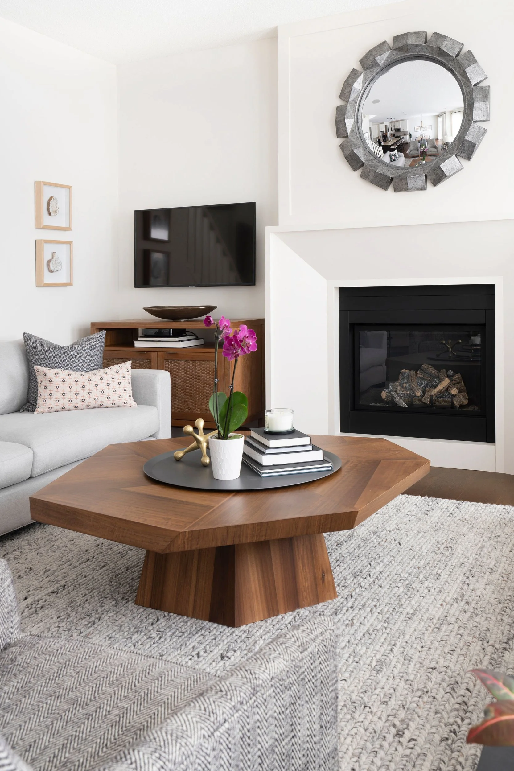 Living room with a wooden coffee table, gray sofa, wall-mounted TV, fireplace, decorative mirror, and orchid plant.