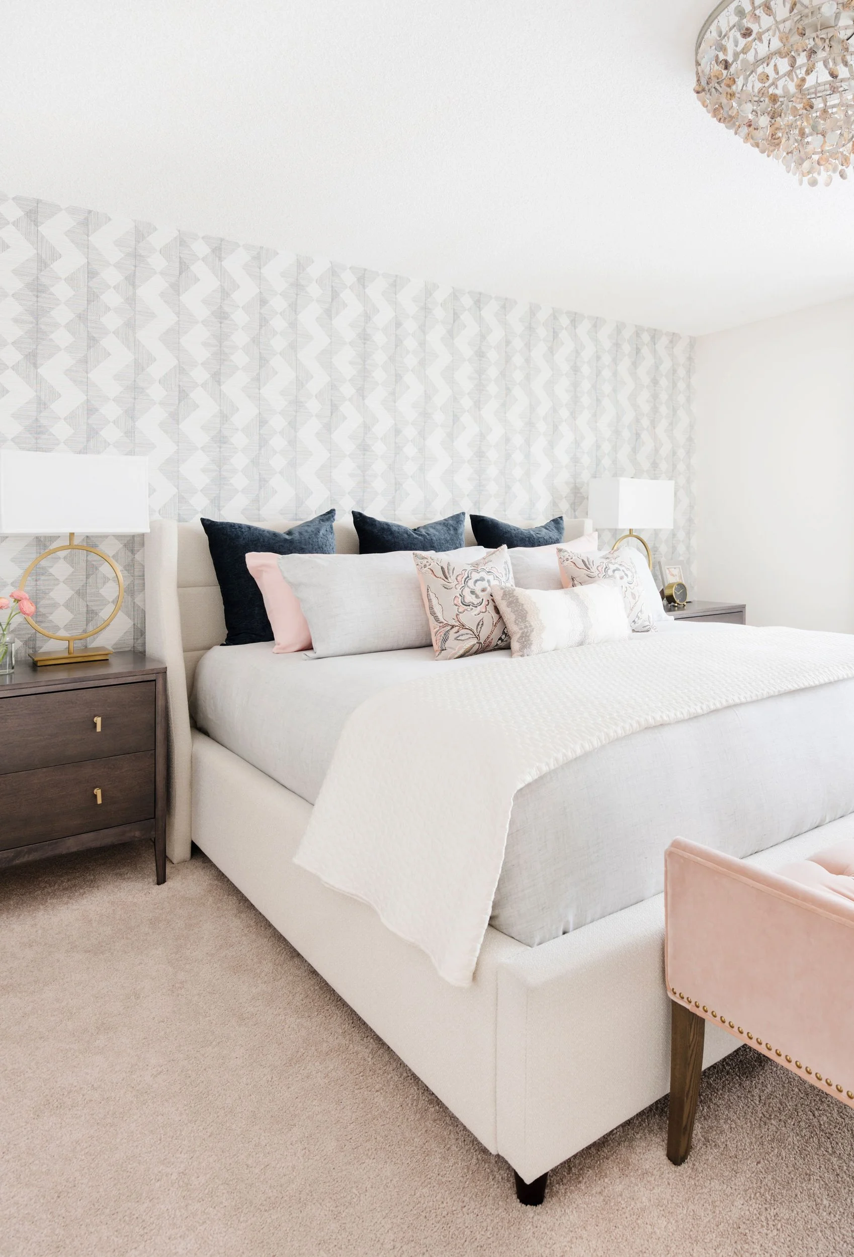 Modern bedroom with geometric wallpaper, upholstered bed with decorative pillows, two dark wood nightstands, and a crystal chandelier.