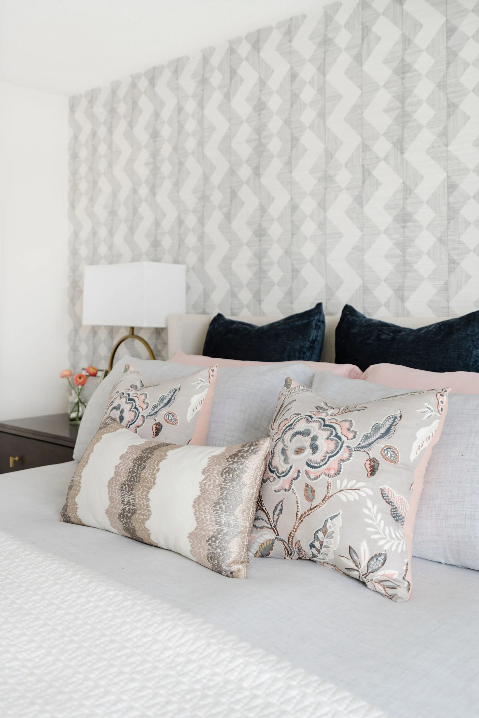 Modern bedroom with decorative floral pillows, a textured gray and white patterned wallpaper, and a bedside table with a lamp and flowers.