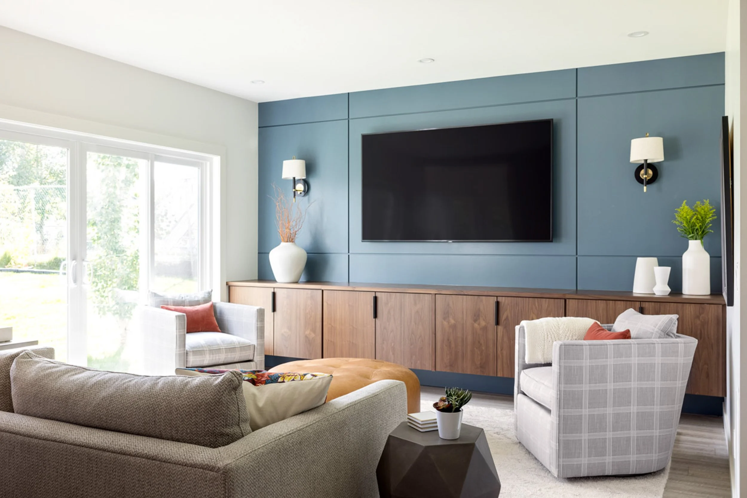 Modern living room with a blue accent wall, large TV, wooden cabinets, and contemporary furniture, including a gray sofa and plaid armchairs with orange cushions.
