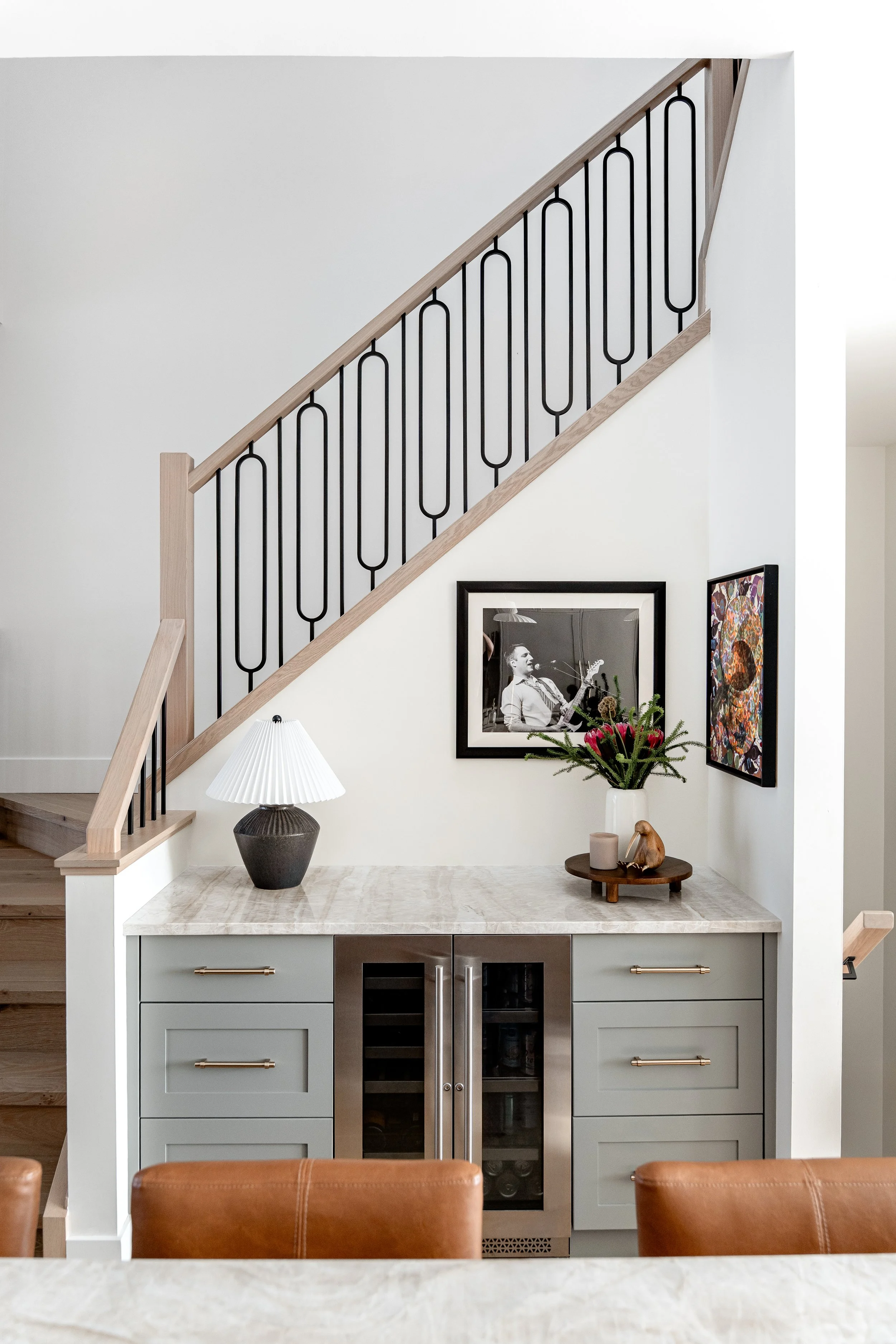 Interior view of a modern kitchen with a marble countertop, light gray cabinets with gold handles, and a built-in wine cooler. Above the countertop, a decorative wall with framed black and white photos and colorful art. A table lamp and a flower vase