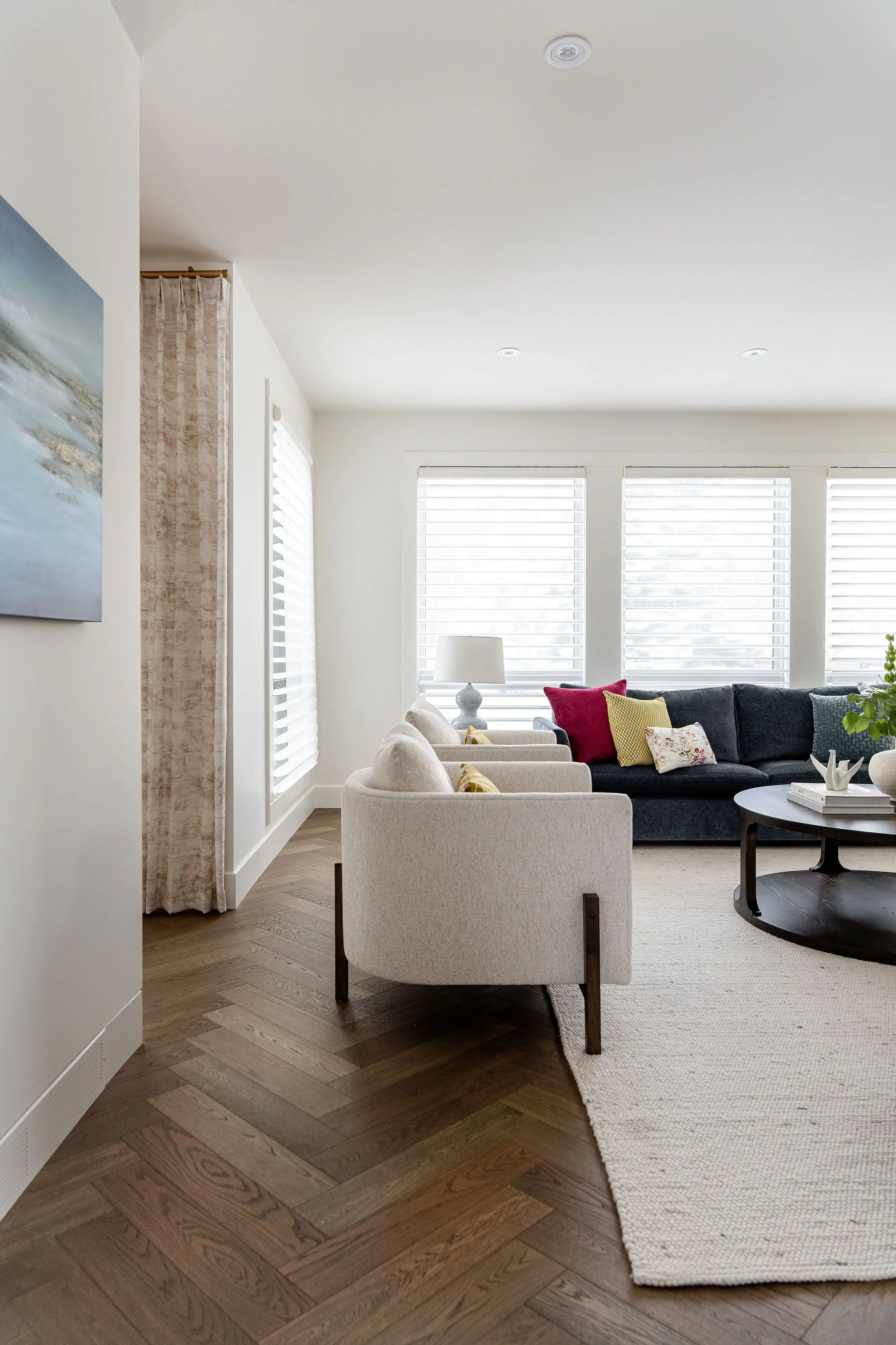 Living room with beige armchairs, a black sofa with colorful pillows, a round black coffee table, and a white lamp on a side table. Large windows with white blinds let in natural light, and a painting is visible on the wall. Light-colored walls and h